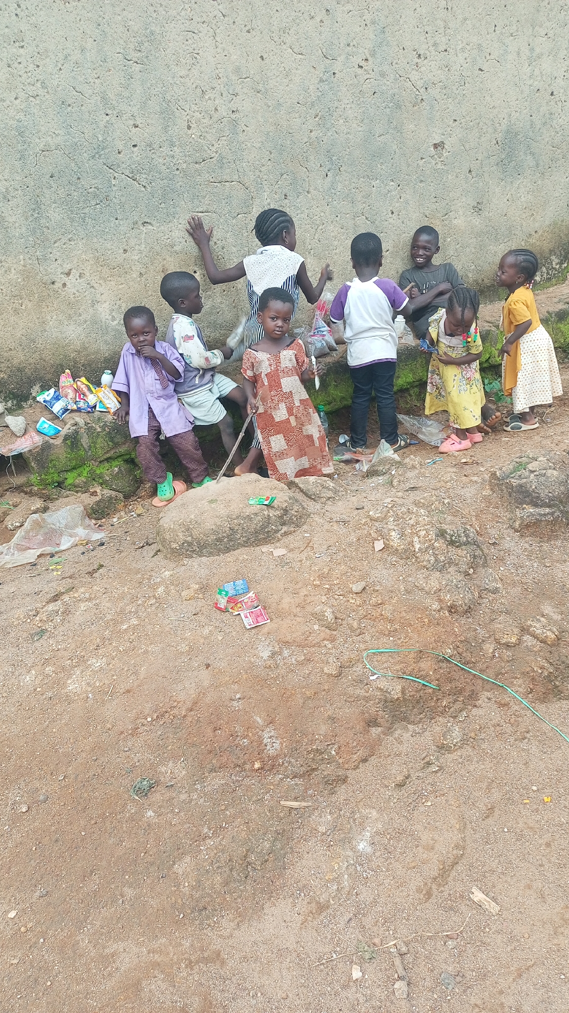 A group of seven young children are gathered near a rough, textured wall in what appears to be an outdoor, informal setting. The children, ranging in age, are dressed in various colorful clothing.
One child in the foreground on the left is wearing a purple tunic and striped pants, with green sandals. Next to them, another child wears a white shirt with a light blue and white striped top. A girl in the center is in a patterned dress. To her right, a child in a white t-shirt with purple accents and dark pants. Another girl to the far right wears a yellow dress with a floral pattern. Behind them, a child in a dark t-shirt is seated, laughing, and another child is partly visible further back.
The ground is made of dirt and rocks, with some scattered debris including plastic bags and what look like small packages of sweets or snacks. Some of these packages are stacked on a rock in the foreground. A green hose or cable is visible on the ground in the lower right of the frame. The wall behind the children is a light, rough material, possibly concrete or plaster, with visible cracks and imperfections.
The lighting suggests it is daytime, with natural light casting shadows. The overall scene conveys a sense of casual interaction and play among the children in what seems to be a rustic or underdeveloped area, potentially in Kagarko South, Nigeria, as suggested by the location context. The children's expressions range from neutral to joyful, with the seated child clearly expressing amusement. No visible text is present in the image.