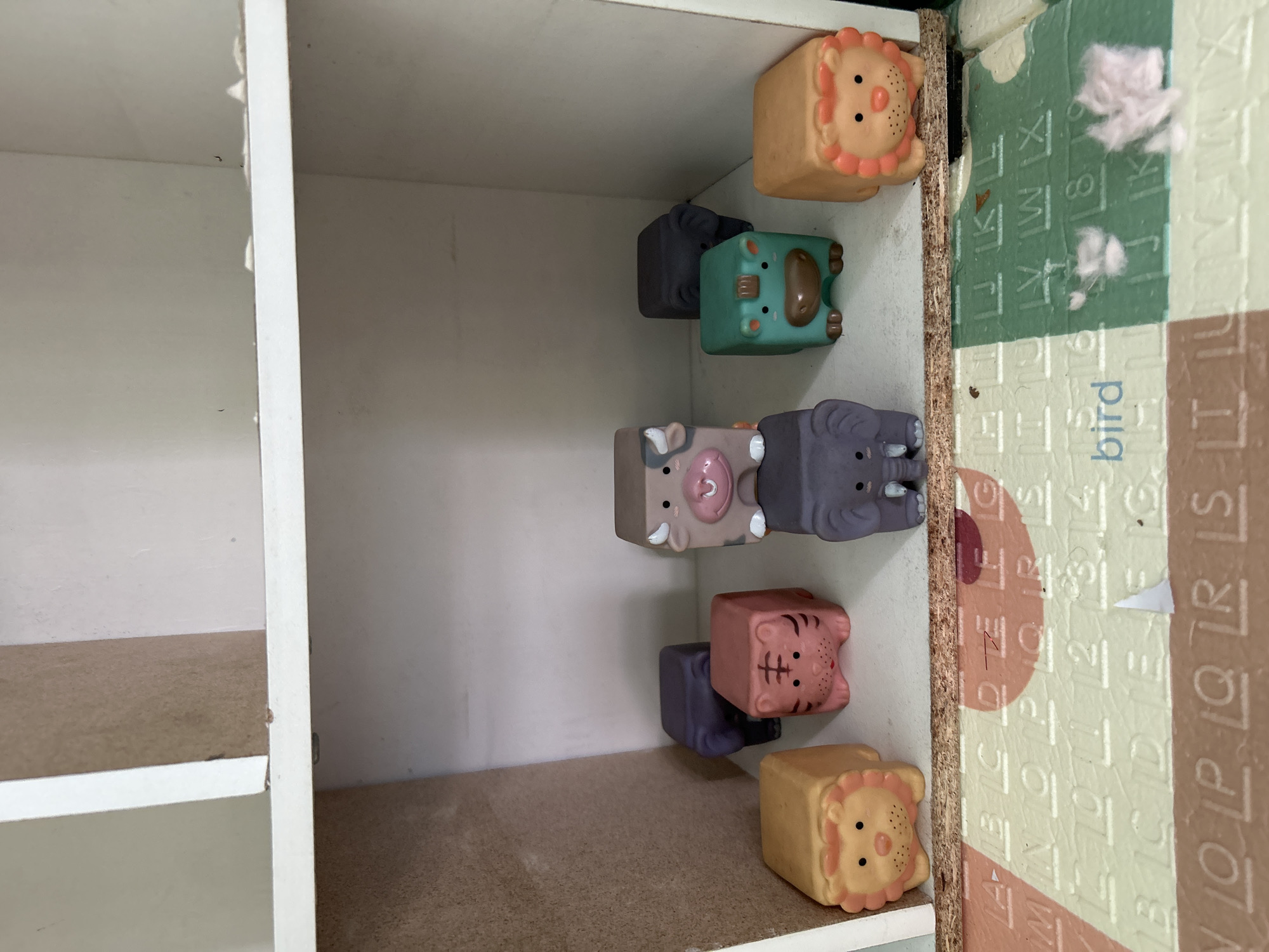In a shelf with a white frame, a collection of eight cubical toy animals are arranged. From left to right, on the bottom row, there is a yellow lion with a prominent mane, a pink tiger with black stripes, a grey elephant, a green hippopotamus, and another yellow lion. Behind the tiger is a dark purple animal, likely an elephant due to its trunk shape, and behind the hippopotamus is a dark grey elephant. In the back row, center, is a light brown cow with pink spots and horns. The toys are placed on a white shelf above a colorful play mat with letters and numbers. The background is a light-colored wall. There are no people or actions depicted. The scene is indoors, possibly in a child's room or playroom.