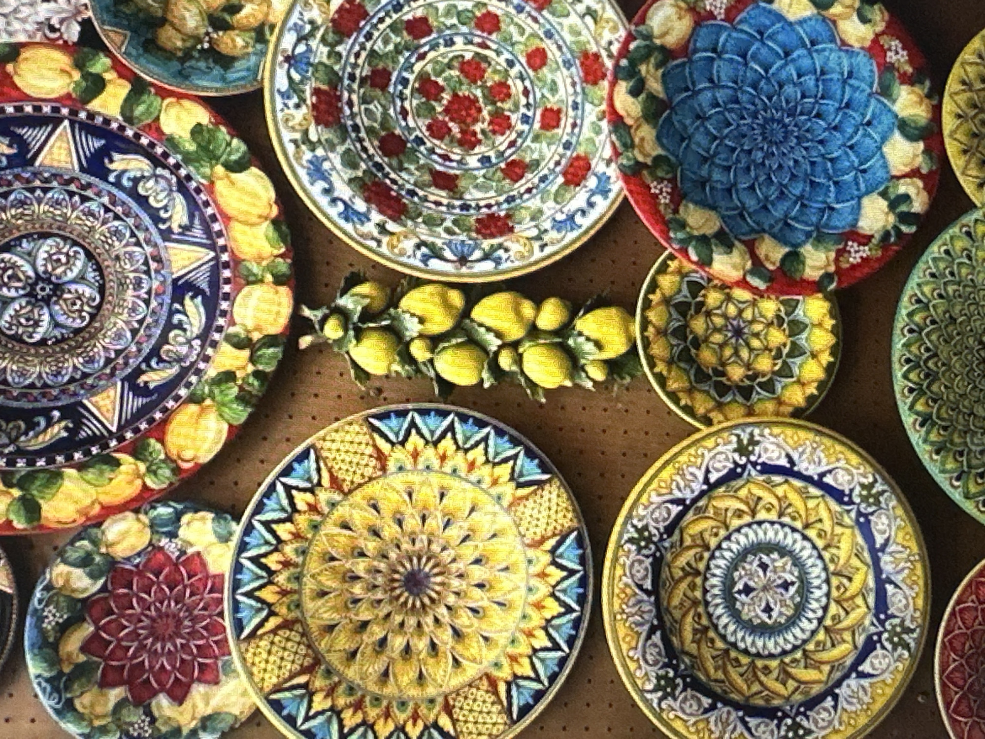 This media file displays a collection of vibrantly decorated ceramic plates, arranged in a top-down view. The plates feature intricate, floral and fruit-themed designs, characteristic of Mediterranean ceramics.
The subjects are the plates themselves, each adorned with a distinct pattern. One plate prominently showcases a large, blue, mandala-like floral design surrounded by yellow fruits and green leaves. Another plate has a central pattern of red flowers on a white background with a blue decorative border. A third plate displays a golden-yellow mandala-style pattern with blue and green accents. Further plates exhibit a deep red floral center with yellow elements, and another with a green, mandala-like design. A garland of yellow lemons and green leaves is placed centrally between some of the plates, adding a unifying theme.
There is no discernible activity or event taking place, as the plates are stationary. The setting appears to be a surface, possibly a table or wall, with a textured, brown background. Notable details include the rich colors and detailed artwork on each plate. There are no visible time of day or weather cues, nor any specific location indicators. The overall impression is one of artistry and decorative appeal. No text is visible in the image.