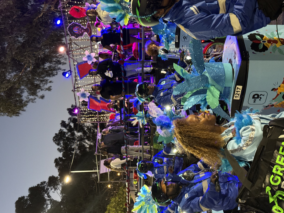 The image displays an outdoor scene at dusk or night, likely during the Cape Town Carnival in Cape Town, South Africa. The foreground features multiple individuals in blue uniforms with reflective elements, dark helmets, and protective eyewear, seated on bicycles or small motorized vehicles. These vehicles are equipped with light blue delivery boxes, branded "Mr D" and numbered "1217" on one visible box. The participants and their vehicles are elaborately decorated with voluminous blue and teal feathered or leaf-like adornments. A woman with light brown, curly hair, wearing a light blue jacket with blue feathered accents, is prominent in the immediate foreground, looking towards the right. Green "GreenDrop" branded bags are also visible.
In the background, a higher platform or float is occupied by several individuals in casual clothing. This structure is brightly illuminated by stage lights emitting blue and pink hues, and decorated with vertical strands of white fairy lights, large red and blue abstract panels, and white and blue floral motifs. A large, dark silhouette of a stylized woman's head is mounted on the far right of this platform. Trees are visible against the dark blue sky in the upper background.
