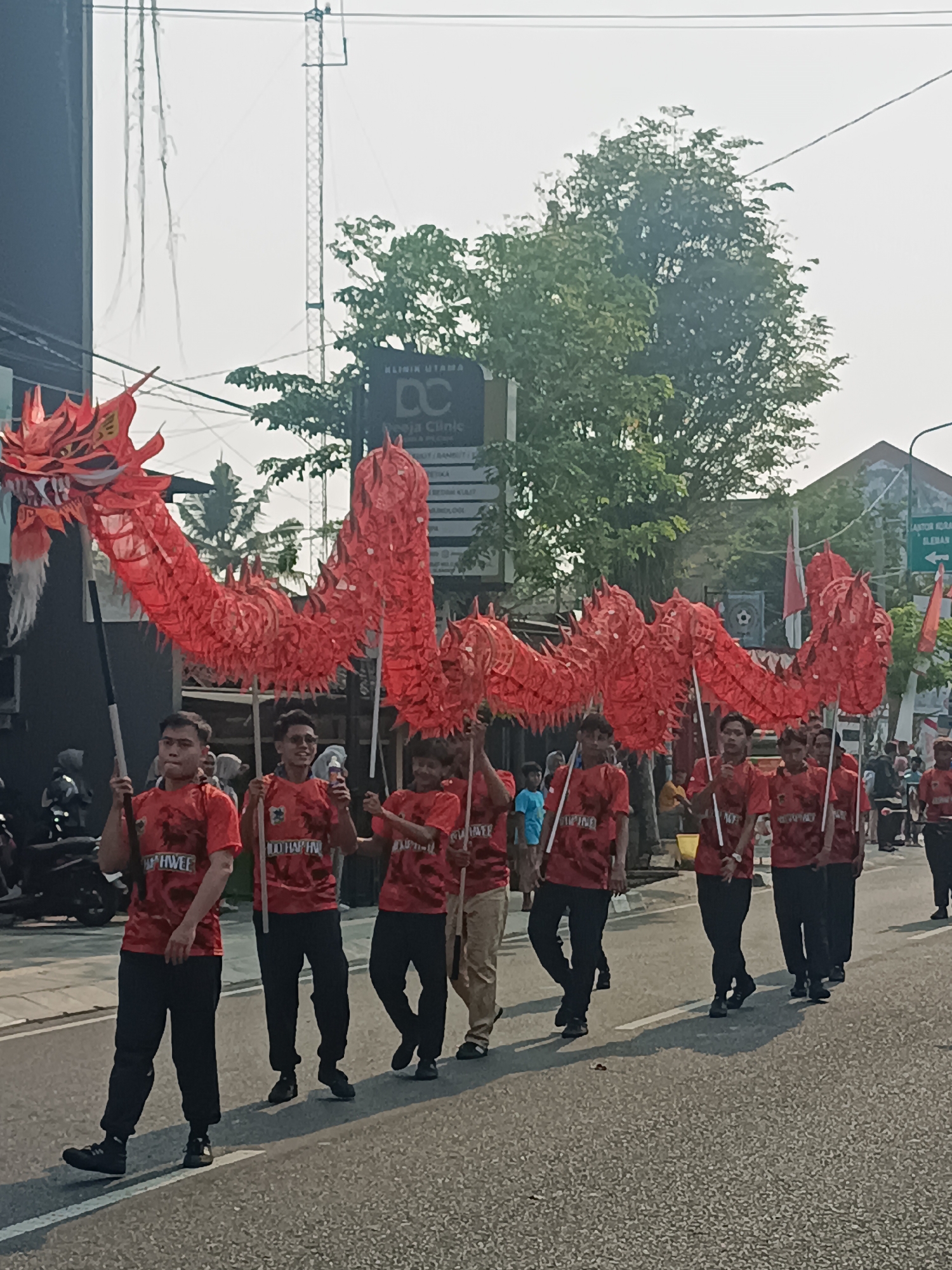 A group of men in matching red shirts and black pants are parading a large, red, segmented dragon float down a street in Sleman, Indonesia. The dragon's head is prominent at the front, held aloft by a man in a black shirt. The dragon extends through the frame, carried by multiple poles held by the parading individuals. The men appear to be walking in unison, their expressions neutral or slightly smiling. The environment suggests a street with buildings and trees lining the sides. A clinic sign with the text "KLINIK UTAMA" is visible in the background, along with a utility pole and overhead wires. The sky is bright and overcast, indicating daytime. Other people and vehicles are present in the background, suggesting a public event or festival.