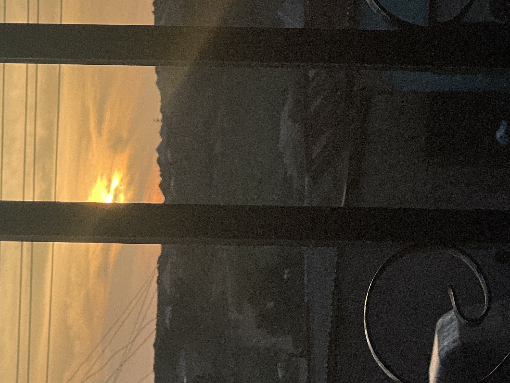 The image captures a sunset viewed through a metal fence in the city of Jos, Nigeria. The sun, a bright orb, is partially obscured by clouds, casting a warm, golden-orange glow across the sky. Rays of sunlight pierce through the clouds, creating a dramatic effect. In the foreground, the dark silhouette of the fence, with its vertical bars and decorative curved elements, frames the scene. Beyond the fence, the cityscape of Jos is visible in silhouette, with hints of buildings and structures under the fading light. Power lines stretch across the horizon, a common sight in urban environments. The overall impression is one of a serene and picturesque sunset over a Nigerian city, with the fence providing a sense of depth and perspective.