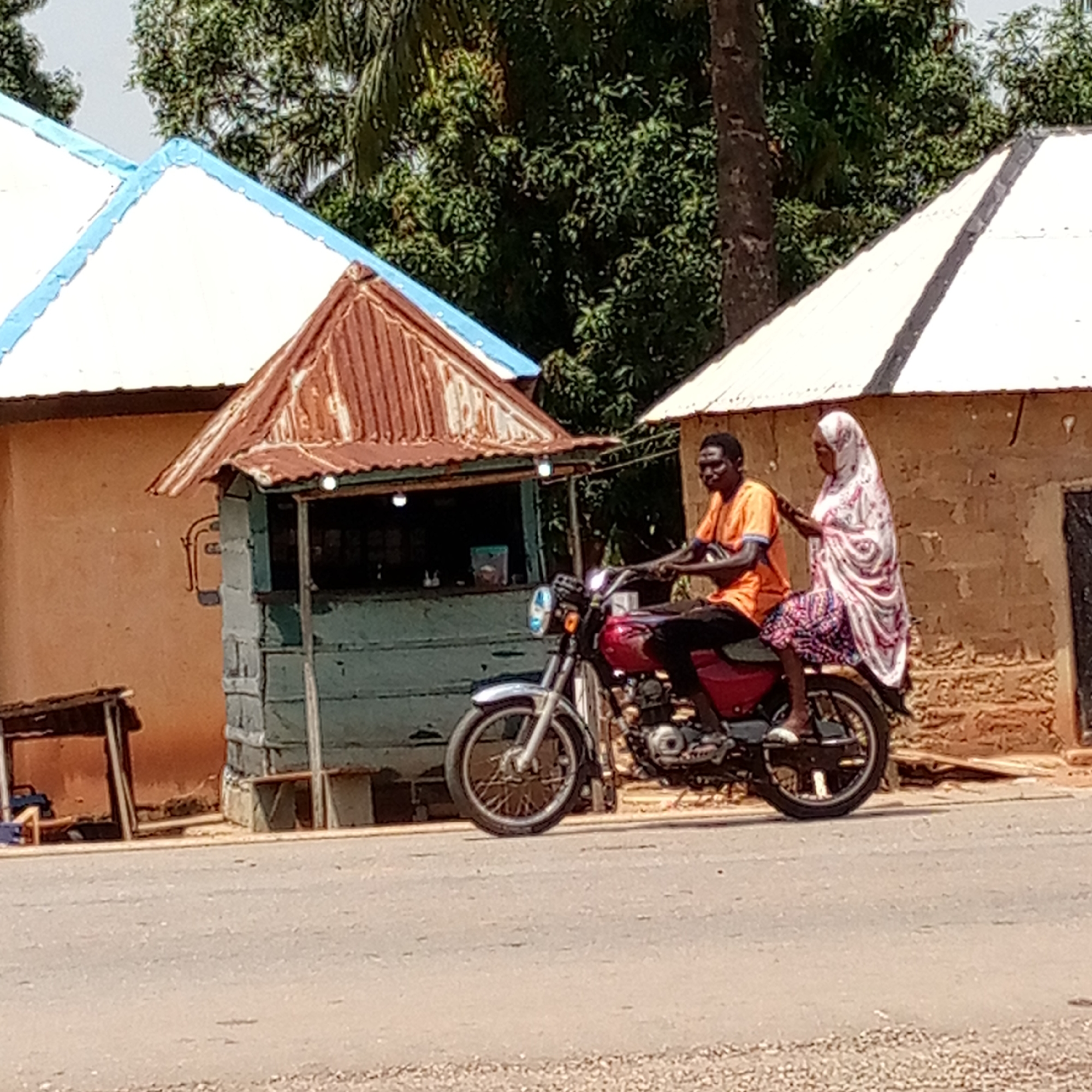 In Mararaba, Nigeria, a man and a woman are riding a red motorcycle on a paved road, with a small blue-and-brown stall and a light-colored building with a blue trim on the roof beside the road. The man is wearing an orange shirt and black pants and is driving the motorcycle. The woman behind him is wearing a white and red head covering and a patterned skirt. Behind the buildings on the side of the road, there are green trees.