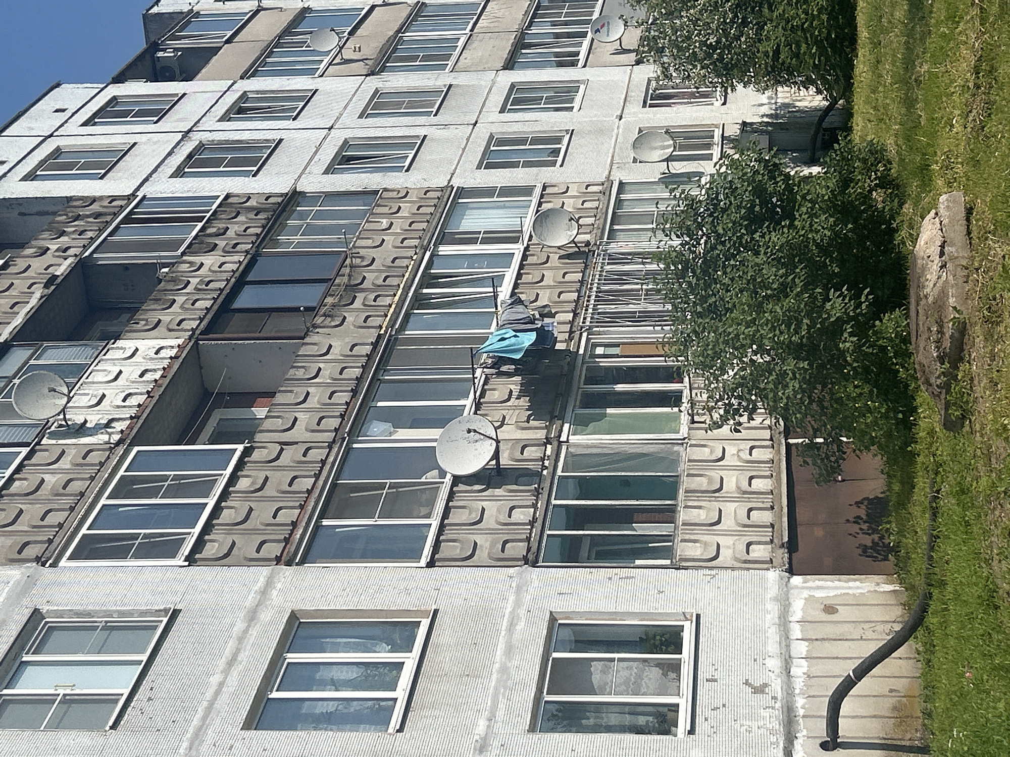 The image shows a multi-story residential building in Posyolok Imeni Morozova, Russia. The building has a white, textured facade and multiple rows of windows and balconies. Some balconies are enclosed with glass, while others are open and feature decorative concrete panels. Satellite dishes are visible on some balconies. Clothes are hanging on a line on one of the balconies. Green bushes and grass are in front of the building. A black, corrugated pipe runs from the base of the building across the lawn. The sky is visible and appears to be blue. The scene is bathed in sunlight, casting shadows on the building and ground.