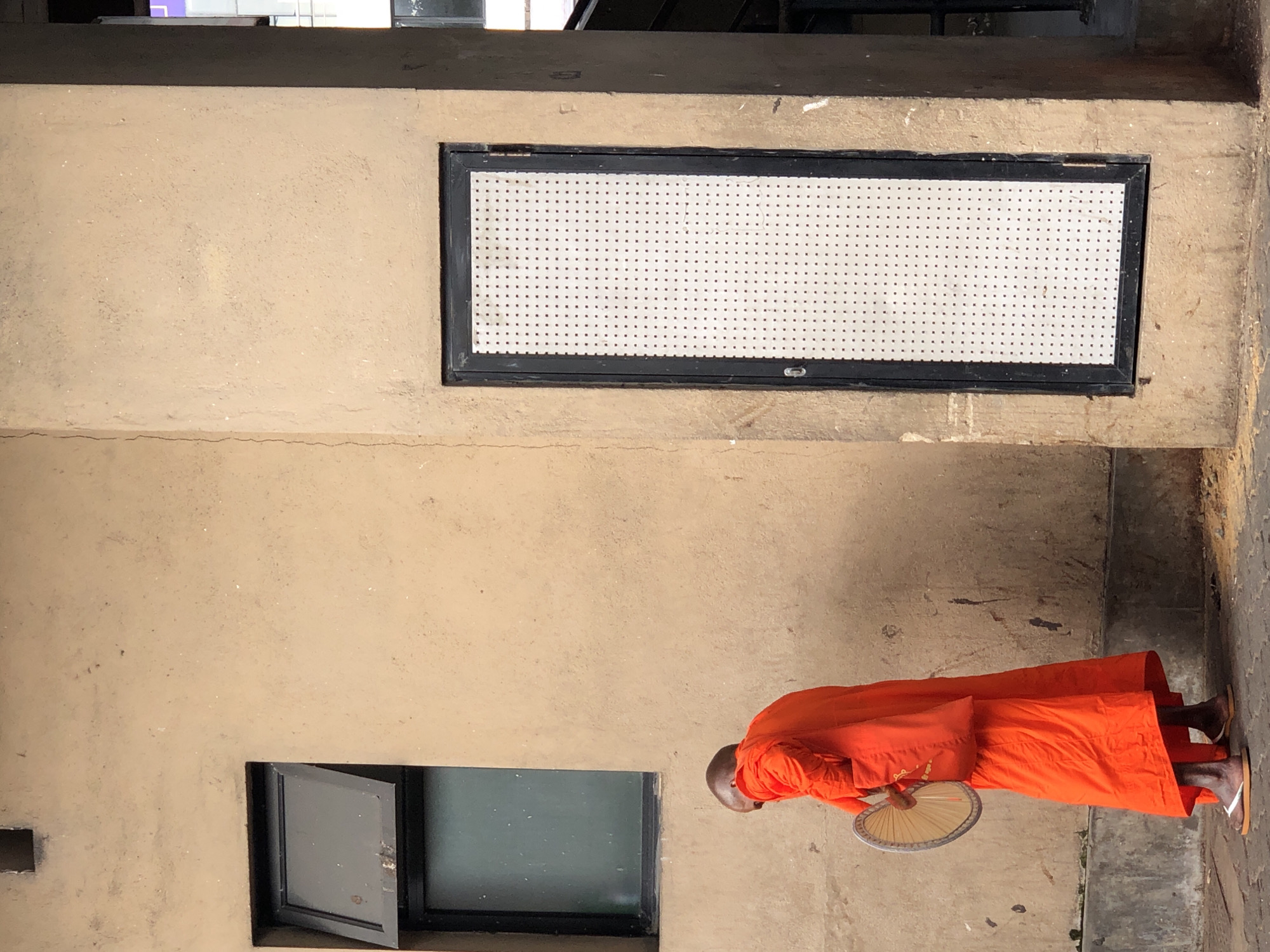 A Buddhist monk, clad in a bright orange robe, stands near a cream-colored building with a dark-framed window and a door with a white perforated panel in Galle, Sri Lanka. The monk is facing away from the camera, holding a circular, fan-like object in his left hand. The building's walls show some texture and a faint crack on the left side. The ground is paved with a rough surface, and there are some scattered debris. A dark concrete curb runs along the bottom of the wall. The lighting suggests an overcast day or shaded area.