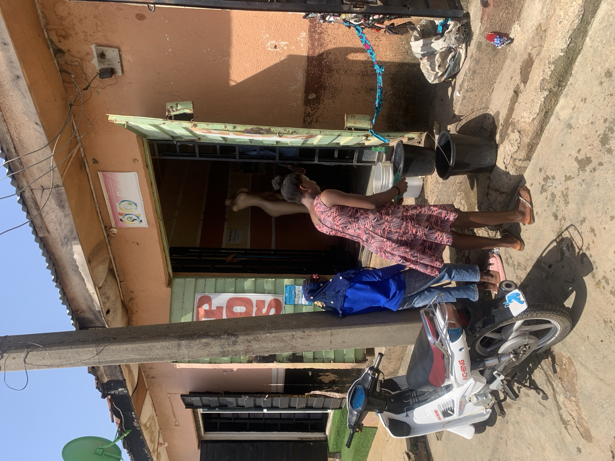 This image depicts a vibrant street scene in Jos, Nigeria, under bright daylight conditions with a clear sky, evidenced by the strong shadows cast on the concrete ground and building facade.
The central subjects are a woman and a child. The woman, wearing a patterned sleeveless dress and flip-flops, stands in front of a building, holding a small white bucket. Her posture suggests she is engaged in a daily activity. To her left, a child in a blue long-sleeved top and blue jeans stands near a white motorbike. A third person's leg is visible inside the building's doorway, where a mannequin leg is also present.
The setting is a building with an orange-brown painted facade and a green metal awning or doorframe. Several black and white plastic buckets are scattered on the concrete ground in front of the building. Visible electrical wiring and a mounted electrical box are high on the wall. A corrugated metal roof is partially visible in the upper left.
Key objects include a white and red motorbike, branded "JINCHENG" and "JC110-18D", with a small blue and white dolphin-like sticker on its rear fender. A green satellite dish is visible in the lower left corner of the frame.
Visible text includes a large, partially obscured red sign that appears to say "GOS" or "DOS". Below it, a smaller white sign reads "P2P PAYS TRANSFER" along with several phone numbers. Another small white sign with a pink and blue graphic is affixed above the doorway. The scene captures a typical moment of daily life in an urban or semi-urban setting.