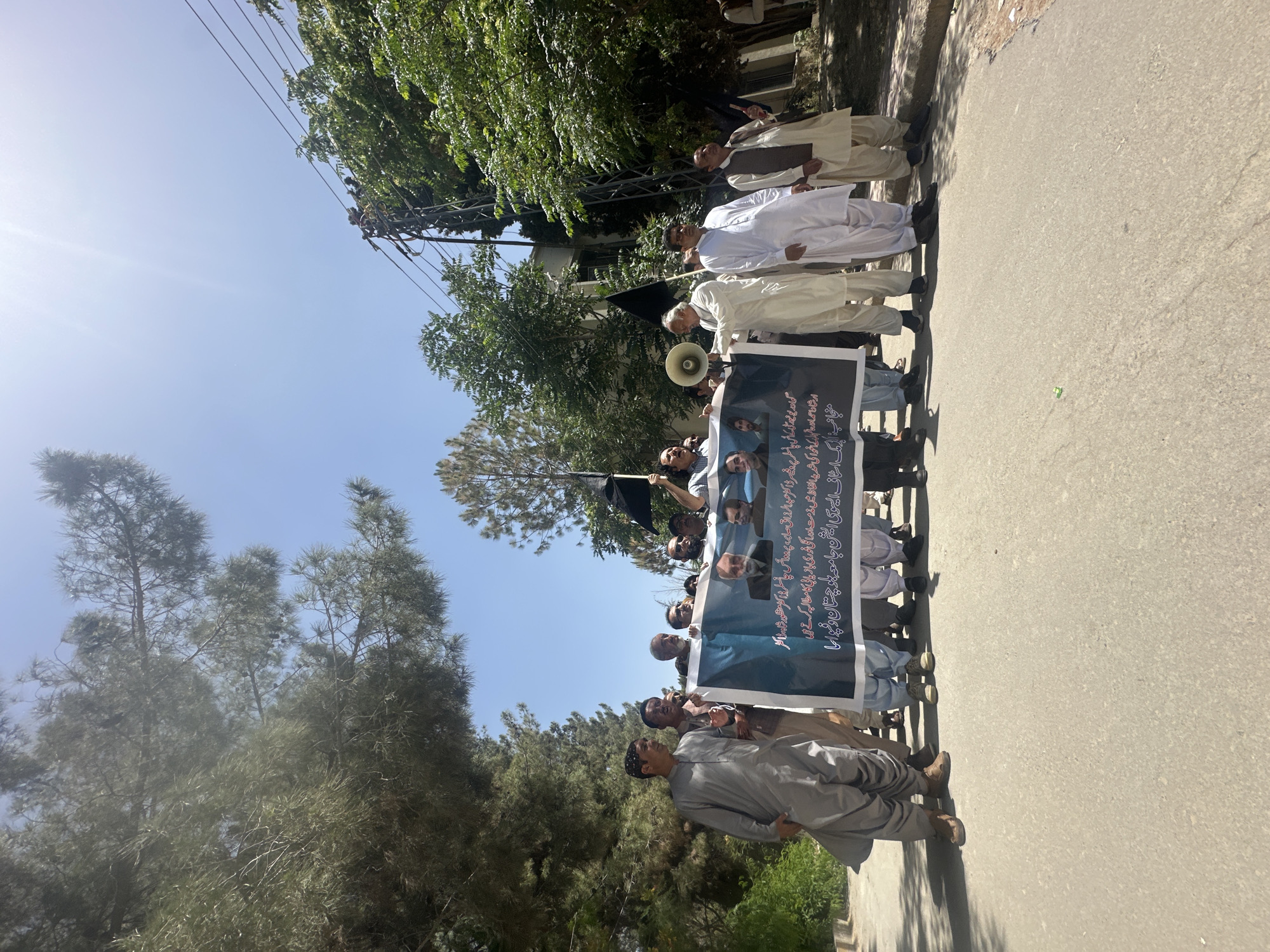 A group of men are standing on a street, holding a banner. They are in Quetta City Tehsil, Pakistan. The banner has text in Urdu and images of people. Some men are wearing traditional Pakistani clothing. One man is holding a megaphone. The sky is bright blue. Trees are visible on both sides of the street. There is a road.