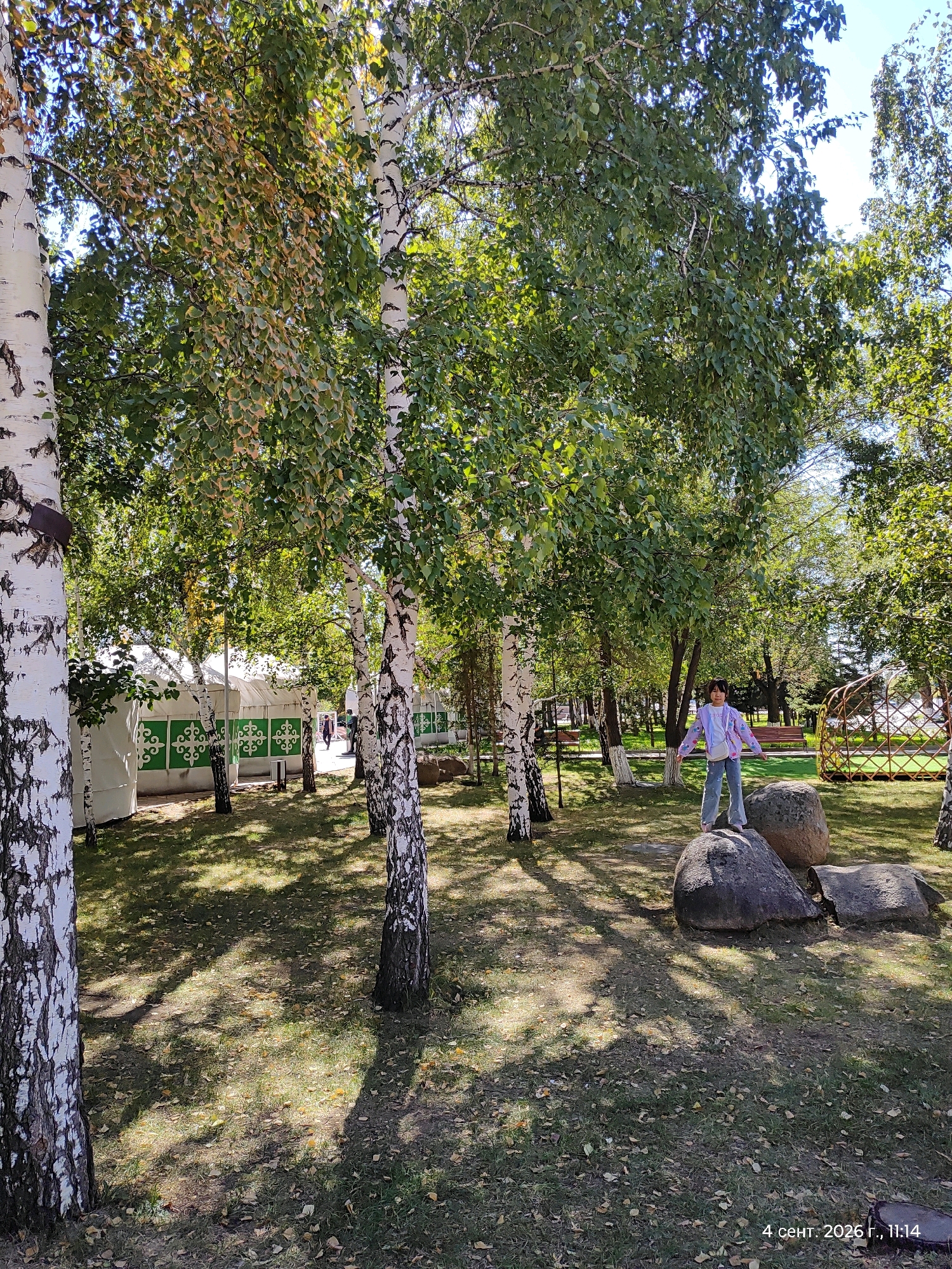 In Astana, Kazakhstan, a young girl stands on large rocks in a park setting. She is wearing a light-colored jacket with a patterned design and blue jeans, her arms outstretched. The park is characterized by numerous birch trees with white bark and green foliage, casting dappled shadows on the grassy ground. In the background, several white tents with green decorative patterns are visible, suggesting a cultural or event space. A traditional-looking gazebo or yurt structure is also present. The time stamp on the image indicates it was taken on September 4, 2026, at 11:14 AM.