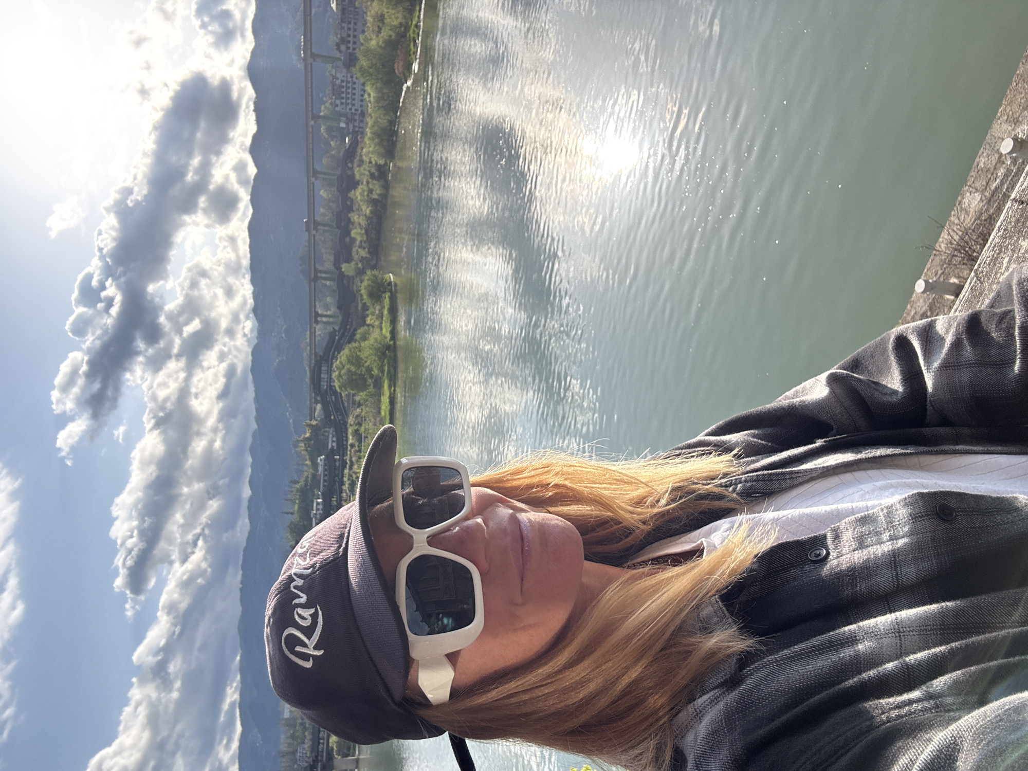 A medium-angle outdoor photograph depicts a person wearing a grey baseball cap with white script reading "Rams", white-framed sunglasses, and a grey and white plaid flannel shirt over a white collared shirt. The person is standing next to a body of water, which likely is Zhuoshui River, given the location details. The river reflects sunlight, causing a shimmering effect on the water's surface. In the background, there is a mountain range partially obscured by clouds. A bridge with a green superstructure spans the river, leading to buildings and trees on the opposite bank. The sky is blue with puffy white clouds on the left side of the frame. The photograph appears to be taken in the city of Zhuoshui, China.