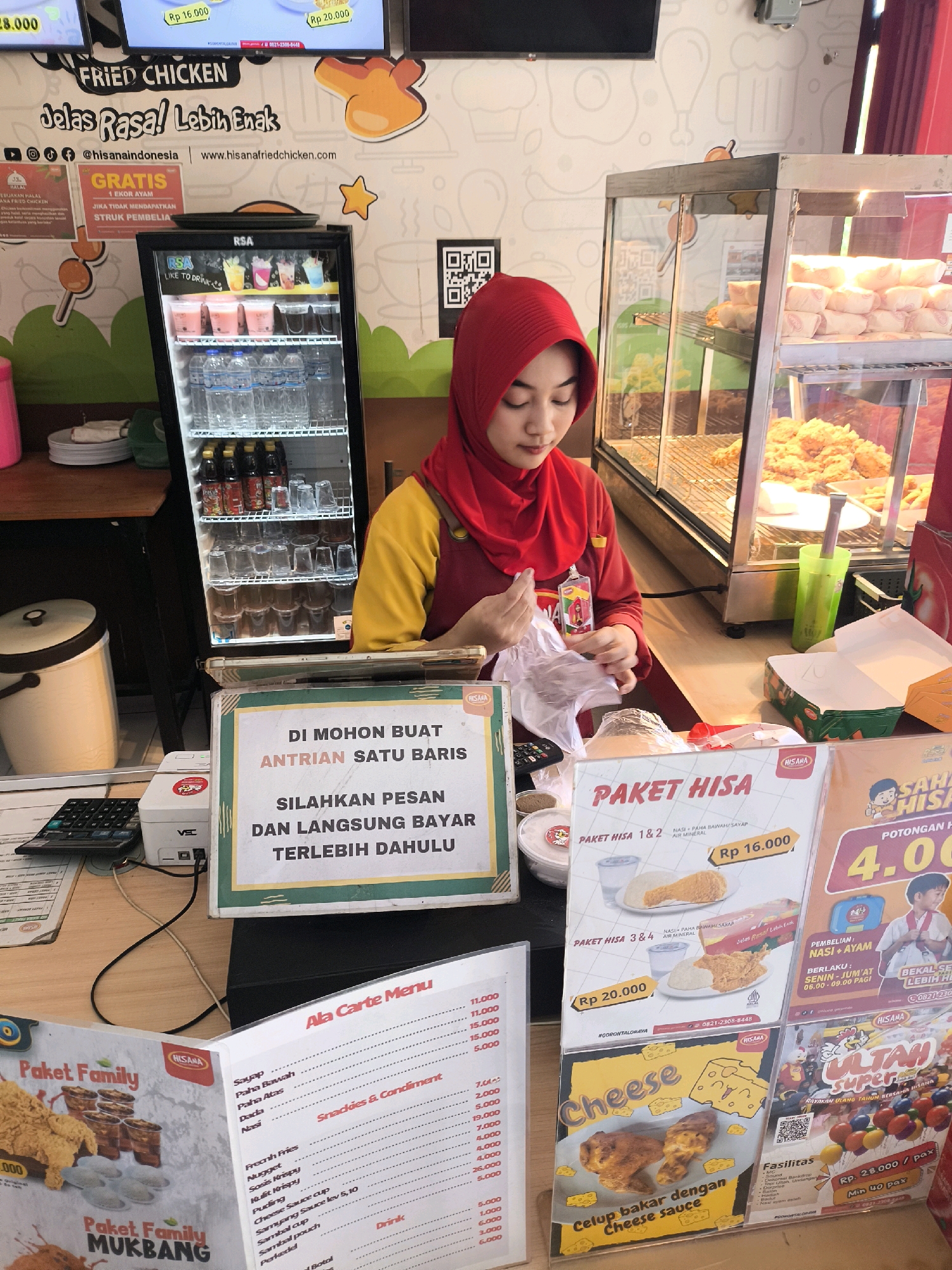 This image depicts the interior of a HISA Fried Chicken restaurant in Gorontalo, Indonesia. A female employee wearing a red hijab and a yellow and red uniform stands behind a counter, holding a plastic bag. She is in the process of packaging food.
In the foreground, there is a calculator, a receipt printer, and a sign that reads "DI MOHON BUAT ANTRIAN SATU BARIS, SILAHKAN PESAN DAN LANGSUNG BAYAR TERLEBIH DAHULU" which translates to "PLEASE FORM A SINGLE LINE, PLEASE ORDER AND PAY FIRST."
Various menu boards and promotional materials are visible. One menu board displays "PAKET HISA" with options and prices. Another highlights "Ala Carte Menu" with prices for various chicken parts, rice, snacks, condiments, and drinks. A "Paket Family" promotion is also shown.
To the left, a refrigerator displays bottled drinks and glasses. Behind the employee, a glass display case contains fried chicken and other food items. A poster with "ULTAH SUPER" and colorful balloons suggests a birthday promotion.
The environment appears to be a casual dining establishment with bright lighting and white walls decorated with chicken-themed illustrations. The overall scene captures a typical busy moment at a fast-food restaurant.