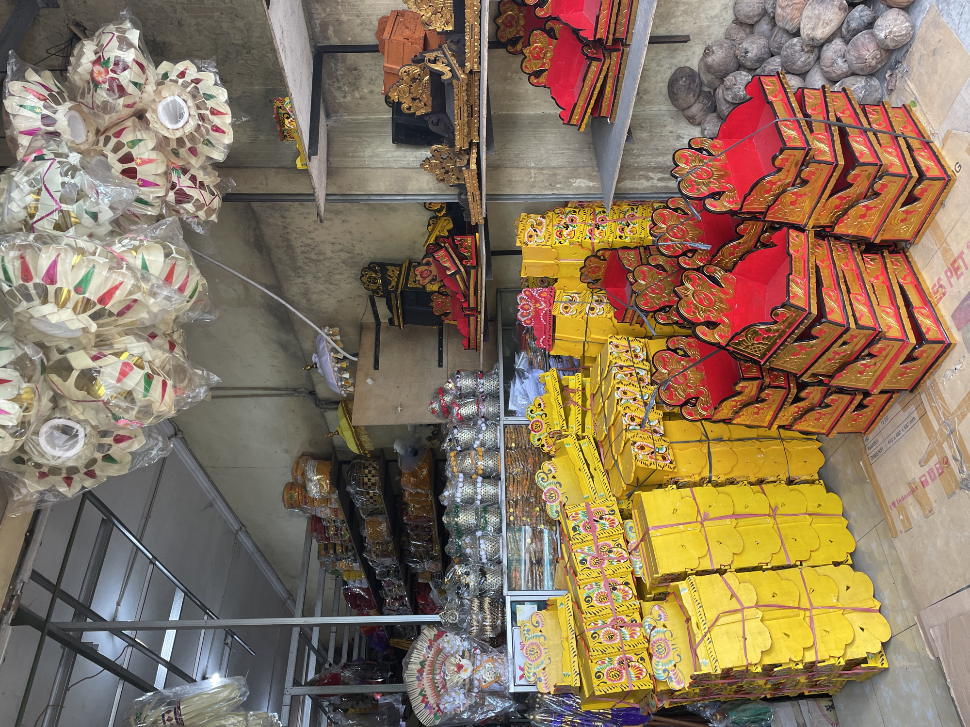 The image captures the interior of what appears to be a storage room or warehouse in Kuta Selatan, Indonesia. Stacks of objects fill the shelves and floor space. There are yellow and red boxes, as well as decorative, white-colored objects resembling lanterns, all likely intended for sale. Additional objects with intricate designs, in black and gold tones, are also present. Some items are wrapped in plastic. The surroundings are utilitarian with metal shelving structures. The items appear to be related to Balinese culture or religious ceremonies.