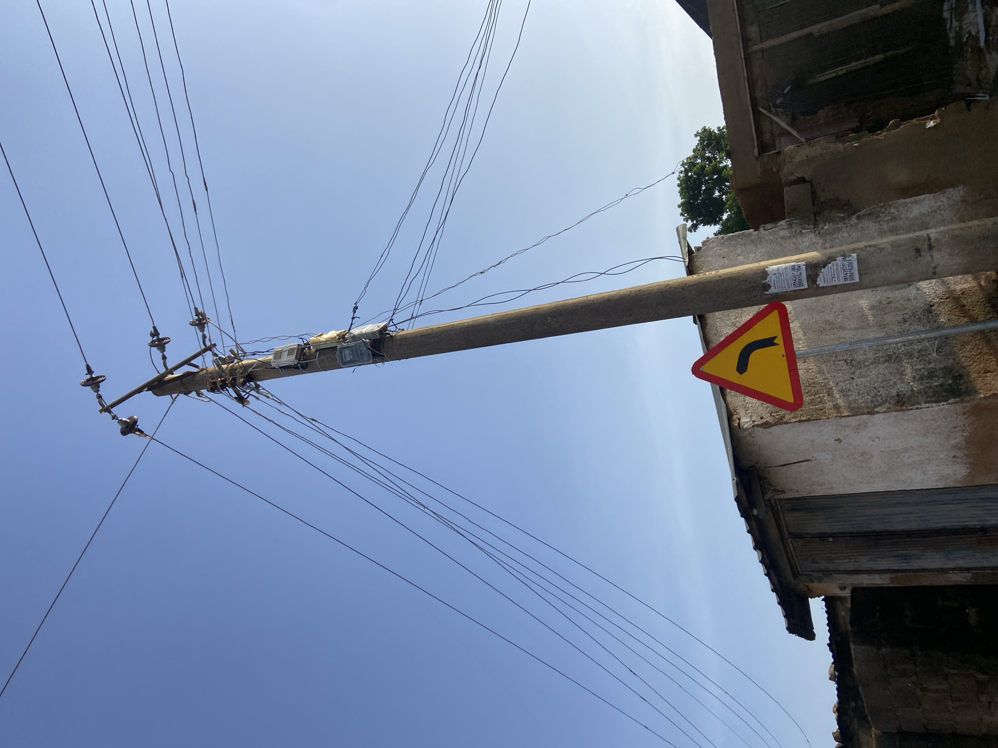 The image depicts an outdoor scene under a clear blue sky. A concrete utility pole, angled slightly, dominates the center of the frame. Numerous electrical wires extend from insulators at the top of the pole and stretch across the sky. Attached to the pole is a yellow, triangular traffic sign with a red border, indicating a left curve. Several electrical meters are mounted on the pole, along with what appear to be advertisements or notices.
To the left of the pole is a dilapidated building with a corrugated metal roof and wooden doors. The walls of the building are rough and discolored. To the right of the pole, another structure with a similar roof is visible, along with a fence constructed of rough-hewn wood. Dense green foliage can be seen behind the structures. The overall impression is of a somewhat neglected urban or semi-urban environment, consistent with certain areas of Jos, Nigeria, where such infrastructure and building conditions are common. There are no people or animals visible in the image.