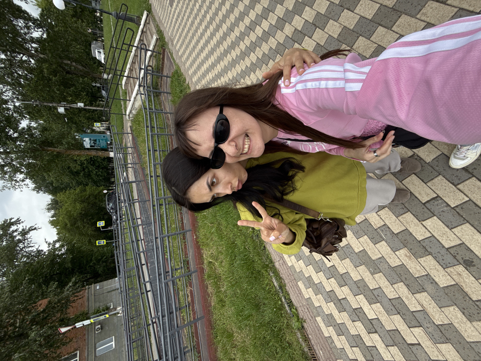This outdoor photograph features two women captured in a selfie-style shot. The woman on the right, in the foreground, wears a pink track jacket with white stripes, dark sunglasses, and has long dark hair. She is smiling broadly at the camera, with her right arm around the shoulder of the other woman. The second woman, positioned to the left, has dark hair and is wearing an olive green hoodie. She is making a peace sign with her right hand and has a slight pout. A brown crossbody bag hangs from her shoulder.
They are standing on a paved walkway made of alternating light gray and dark gray rectangular blocks. To their left, there is a grassy area bordered by a dark metal railing or fence. Beyond the railing, several trees and a street are visible. A white car can be seen on the street, along with two yellow pedestrian crossing signs. In the distance, parts of a building with windows and an air conditioning unit are visible. The sky appears overcast, suggesting it is daytime on a cloudy day. Both women seem cheerful and engaged in the moment. The scene depicts an urban setting, possibly a park or sidewalk area in Votkinsk, Russia.