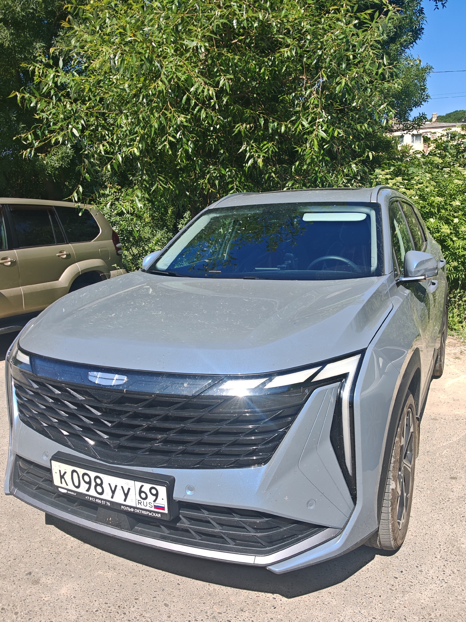 A grey Geely Coolray car is parked on a paved area in front of dense green trees. The car faces forward, slightly angled to the right, showing its front grille, headlights, and license plate. The license plate reads "K098YY 69 RUS". Behind the Geely, another, older model SUV in a sandy brown color is partially visible. The environment suggests a sunny day, possibly in a residential area or a parking lot with ample vegetation. The specific location identified as Varvarovka, Russia, implies a regional context for the scene. There are no people visible in the image.