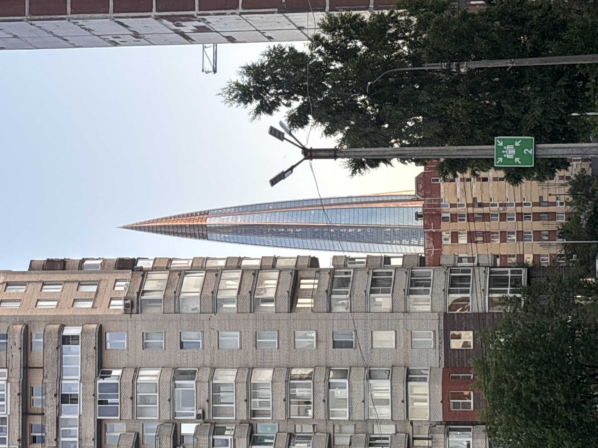 This is a vertical, eye-level, full shot of several buildings with a clear sky in the background. On the left is a tall, light brown brick apartment building with many windows and balconies. To its right is a very tall, modern skyscraper with a pointed, triangular top and a glass facade that reflects the light. In front of the skyscraper and slightly to its right is a large, dark green tree. To the right of the tree is a tall, light gray utility pole with several streetlights attached to it, and a green sign depicting people and arrows pointing inwards, with the number '2' below them. Further to the right is another building with a reddish-brown brick facade. The scene appears to be in an urban environment.