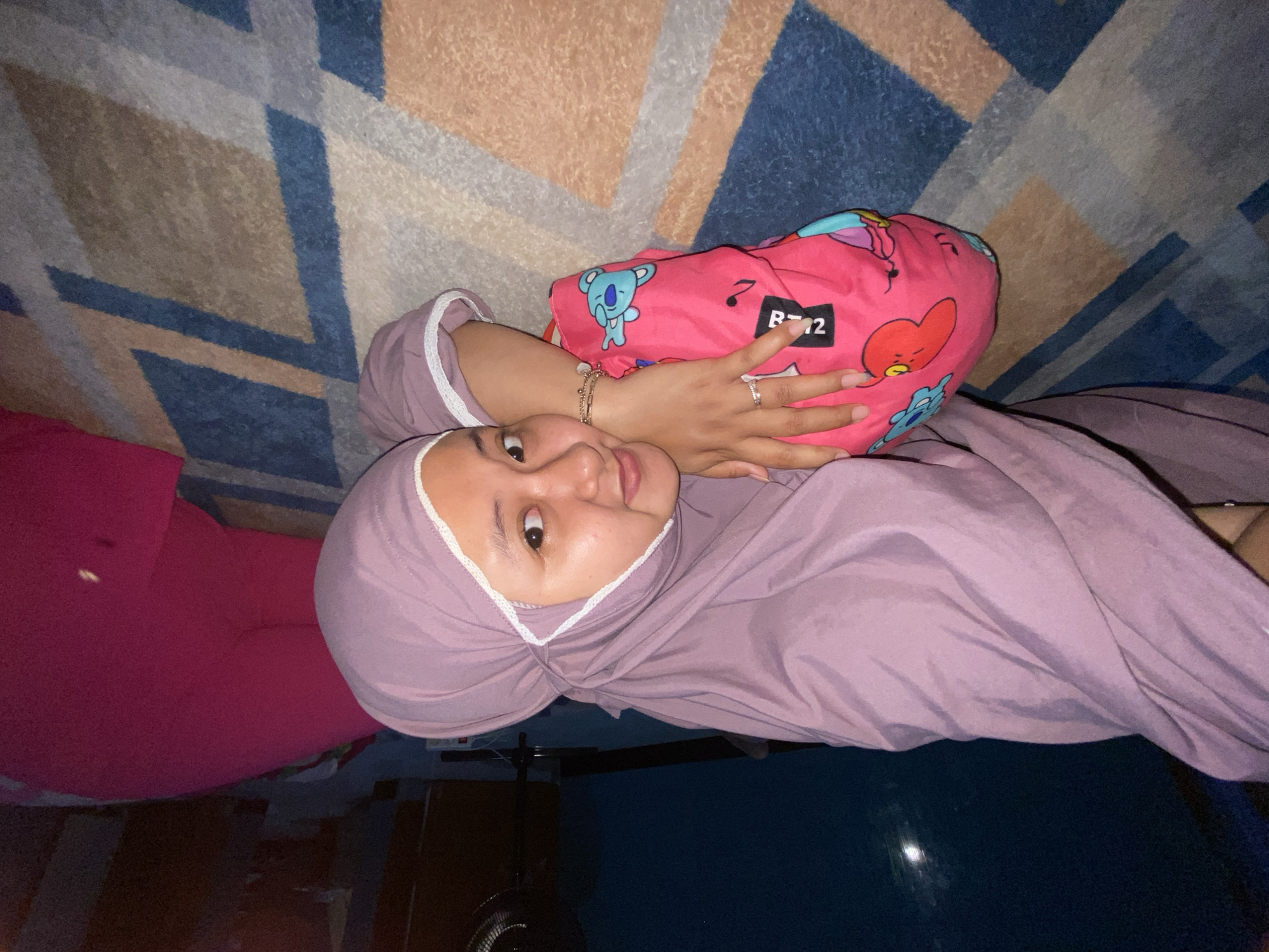 An indoor shot captures a young person, likely female, lying horizontally and looking towards the camera with a gentle smile. She is wearing a light purple hijab with white lace trim along the edges and matching long-sleeved clothing. Her head rests on her right arm, and her left arm is bent, holding a bright pink pillow or cushion adorned with colorful cartoon characters, specifically from BT21. Visible characters include Cooky, Tata, and Koya. A black tag on the pillow clearly shows the text "BT21" and "RJ".
The person's right hand features a silver-toned ring on the ring finger, and her left wrist has a gold-toned bracelet. She is positioned on a patterned rug or carpet with geometric designs in shades of blue, brown, beige, and grey. To the left, a portion of a vivid pink garment is visible, suggesting another piece of clothing or fabric. The lighting appears soft and artificial, characteristic of an indoor setting, possibly during the evening or night. The overall mood conveyed is one of relaxation and contentment.