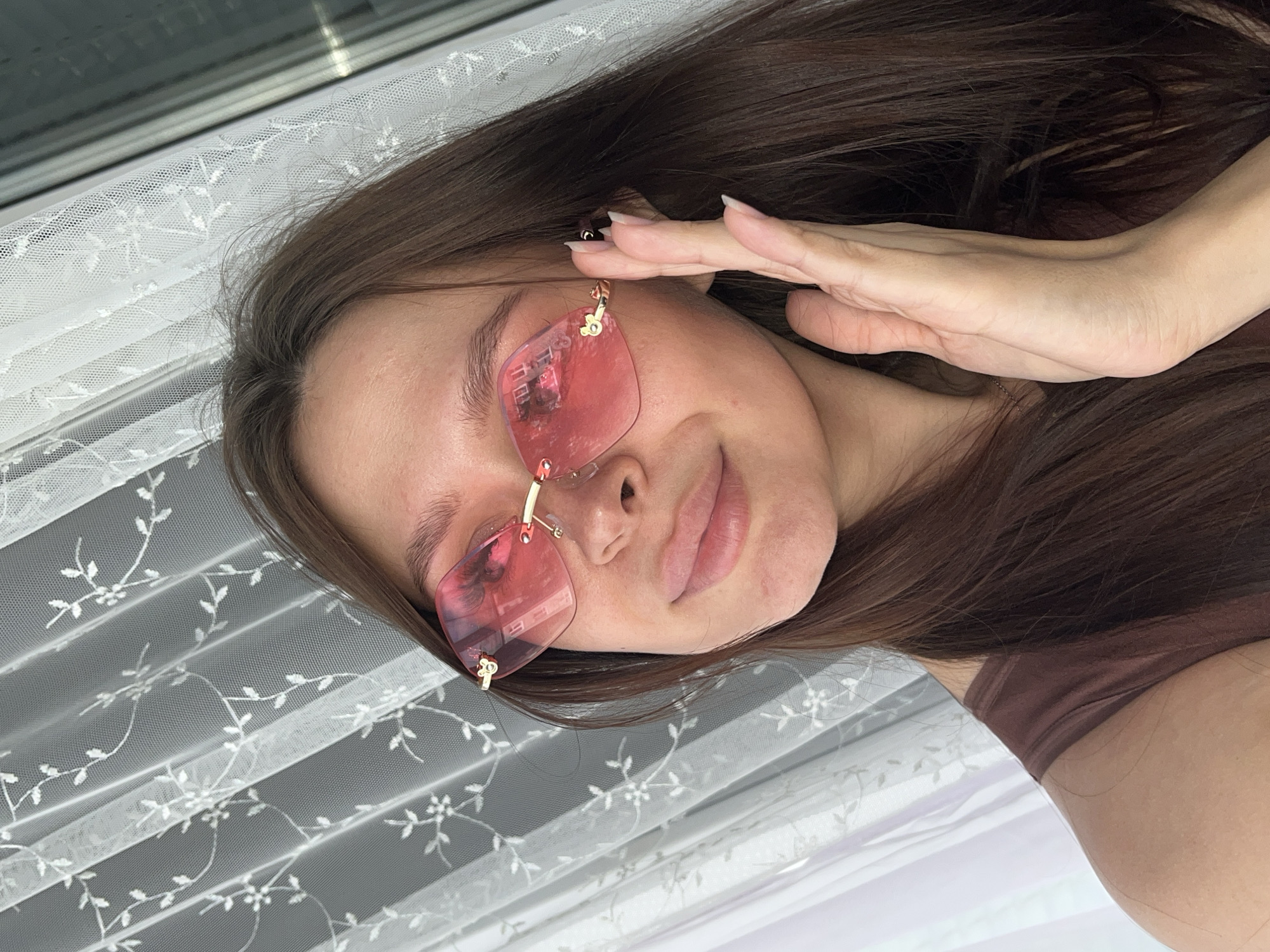 The media file shows a close-up, rotated shot of a young woman with long, dark brown hair. She is wearing pink, rimless sunglasses with gold accents. Her right hand is raised, touching her forehead and the side of her face, with manicured nails visible. The background consists of a white, sheer lace curtain with a floral pattern, which is pleated and draped. Behind the curtain, a window or blinds are visible, suggesting an indoor setting. The lighting appears to be natural, possibly from the window, creating a soft glow. The woman's expression is relaxed and slightly playful, with her lips parted. The overall mood is casual and intimate. There is no visible text in the image. The scene is likely set in Kazan, Russia, based on the provided location context.