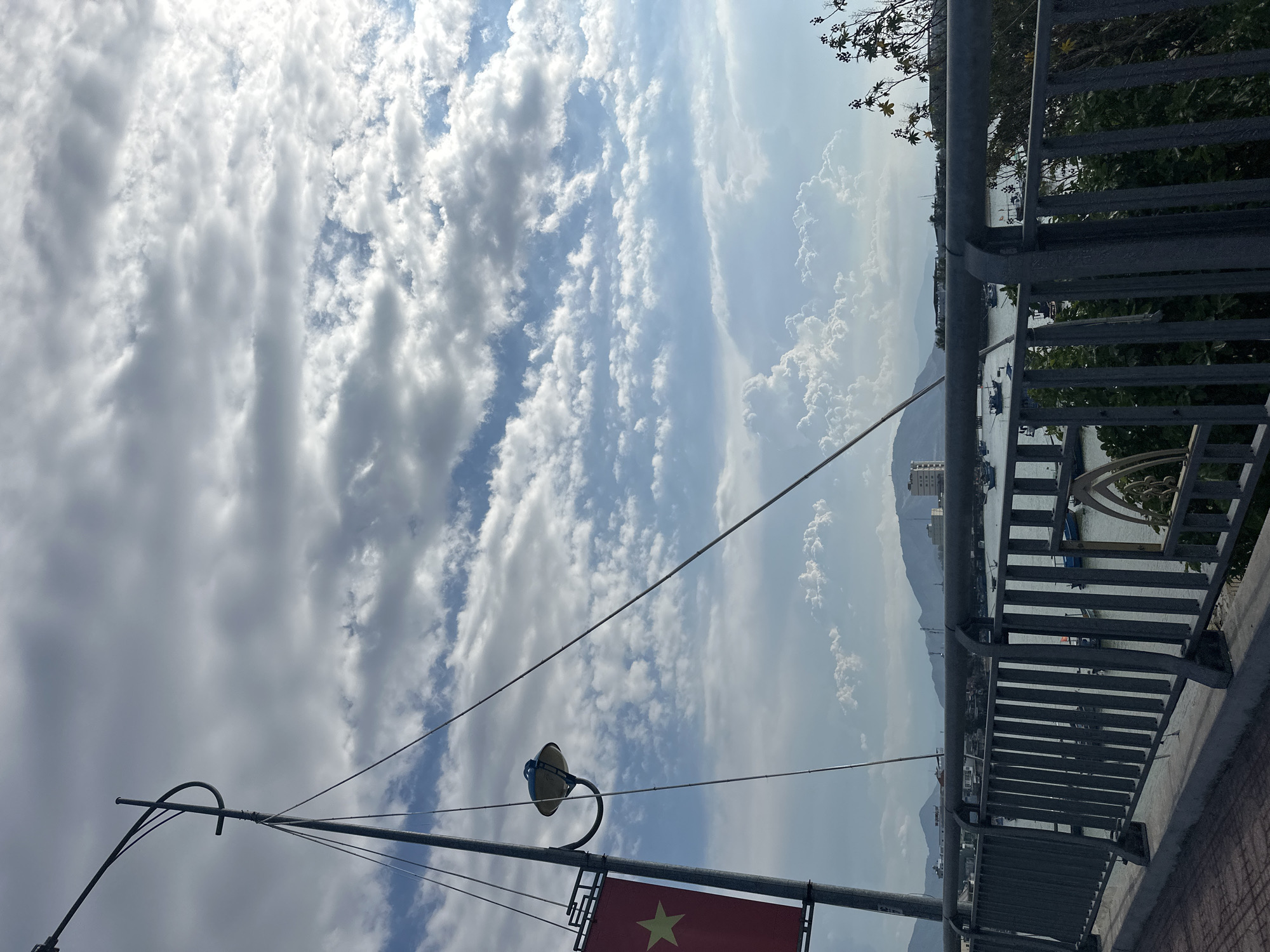 The image depicts a scene in Bac Nha Trang Ward, Vietnam. The sky is predominantly covered with clouds, with patches of blue visible. In the foreground, there is a lamppost and a Vietnamese flag. A metal railing runs along the right side of the frame, overlooking water with boats. Mountains are visible in the distance.