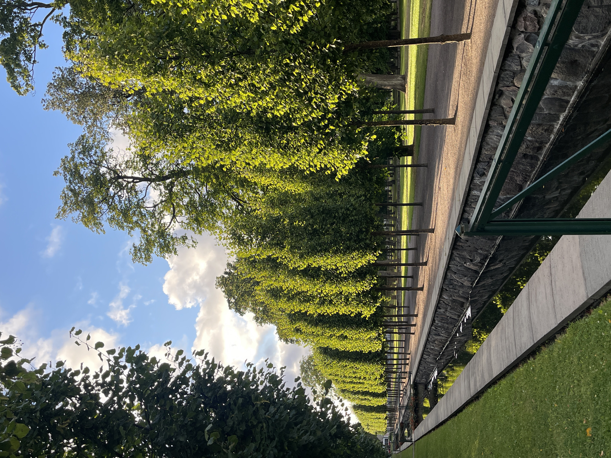 A symmetrical, tree-lined pathway runs alongside a narrow canal in a park. On the left, a dense line of dark green trees forms a wall. The canal, edged with stone on the right and concrete on the left, reflects the sky and trees. Beyond the canal, a row of slender, evenly spaced trees with bright green leaves is illuminated by sunlight, casting long shadows on the gravel path. In the distance, more trees and patches of grass are visible under a partly cloudy blue sky. The scene appears serene and orderly, typical of a formal garden or park.