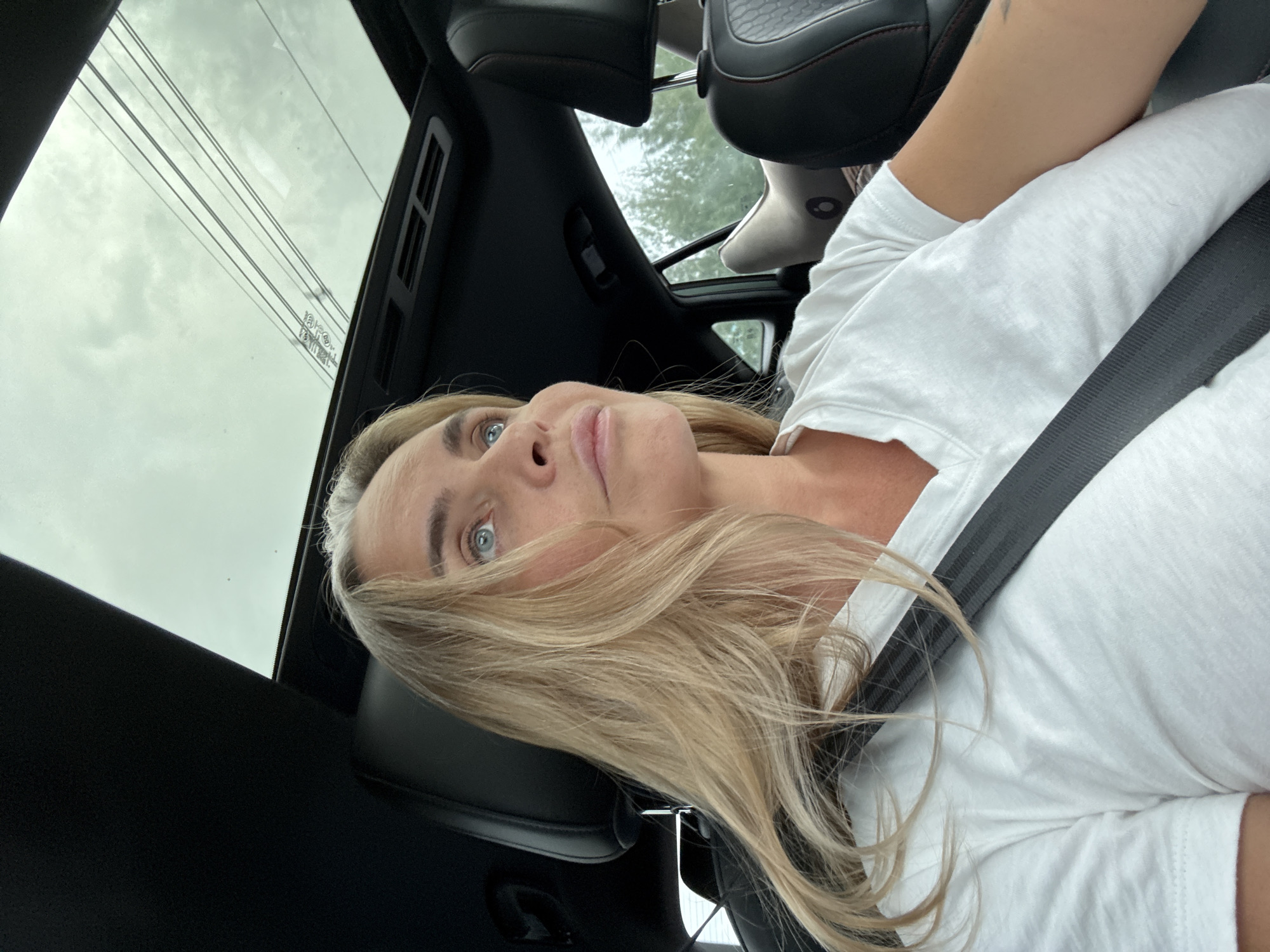 A woman with blonde hair and blue eyes is sitting in the driver's seat of a car, looking upwards and to the right. She is wearing a white V-neck t-shirt and a black seatbelt. The car has a sunroof, which is open, revealing a cloudy sky and power lines. The interior of the car appears to be black leather. The background outside the car's window is blurred, showing trees and foliage, suggesting a location outside of a dense urban area, possibly in or around Ban Nong Bua, Thailand.