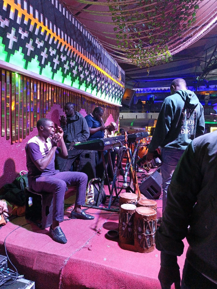 A live band performs on an elevated stage in a colorful venue. A seated man in purple plays percussion while two others play keyboards and another instrument. A man in a hooded sweatshirt stands nearby. The venue features ornate designs, including lit decorative elements and floral accents hanging from the ceiling. The lighting is vibrant, creating a lively atmosphere.