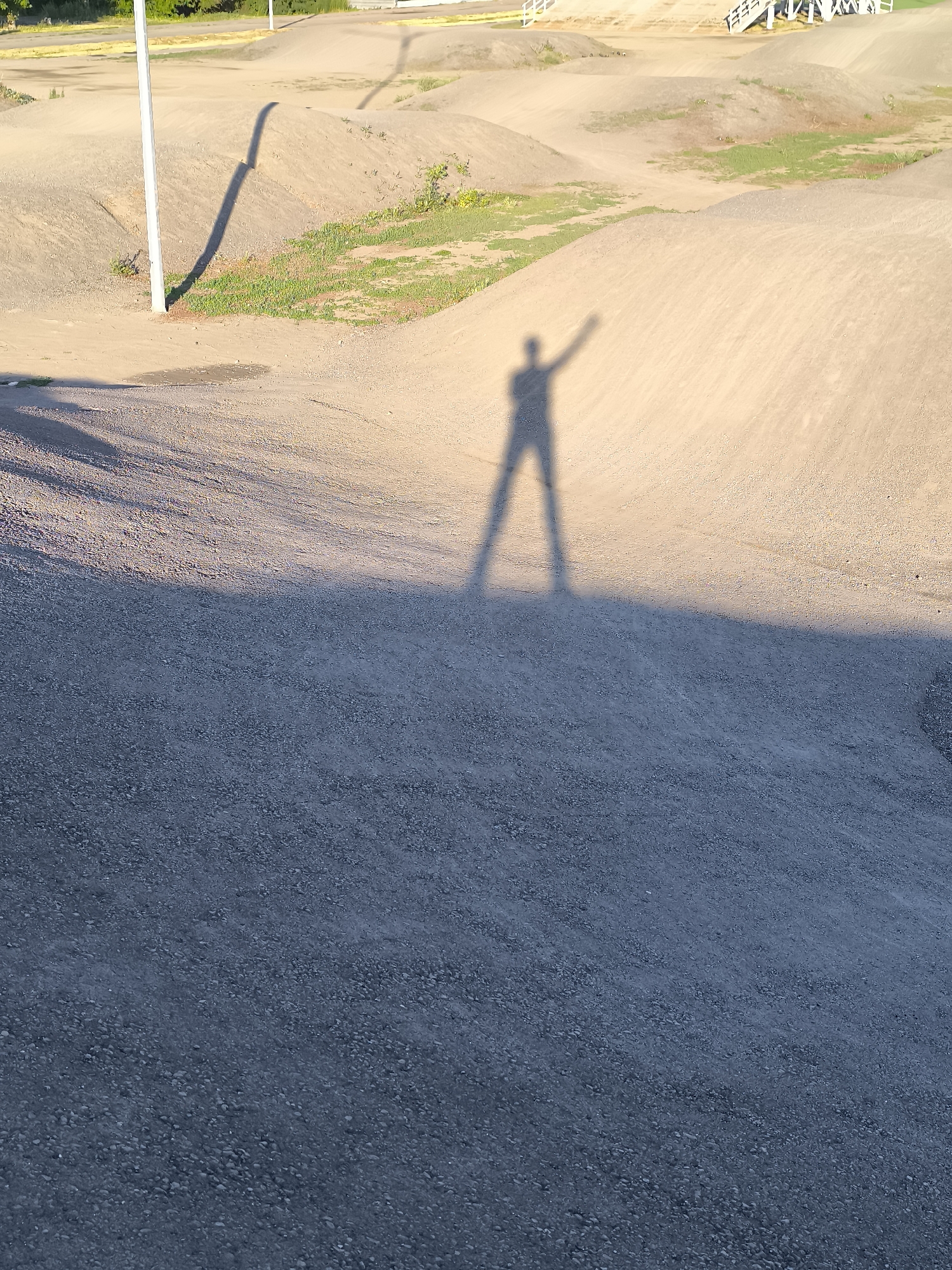 The image shows a shadow of a person standing on a sandy, undulating terrain, possibly a bike park or a natural landscape with hills. The person's shadow is cast to the left, with one arm raised as if waving or celebrating. The ground is composed of fine gravel in the foreground and lighter sand on the raised mounds in the background. A tall, thin, white pole with a shadow stretching across the ground is visible to the left of the frame. In the distance, there are hints of trees and structures, suggesting an outdoor recreational area within a city. The lighting indicates it is either early morning or late afternoon. The scene does not contain any identifiable landmarks that would pinpoint it to the city administration of Qarağandı, Kazakhstan.