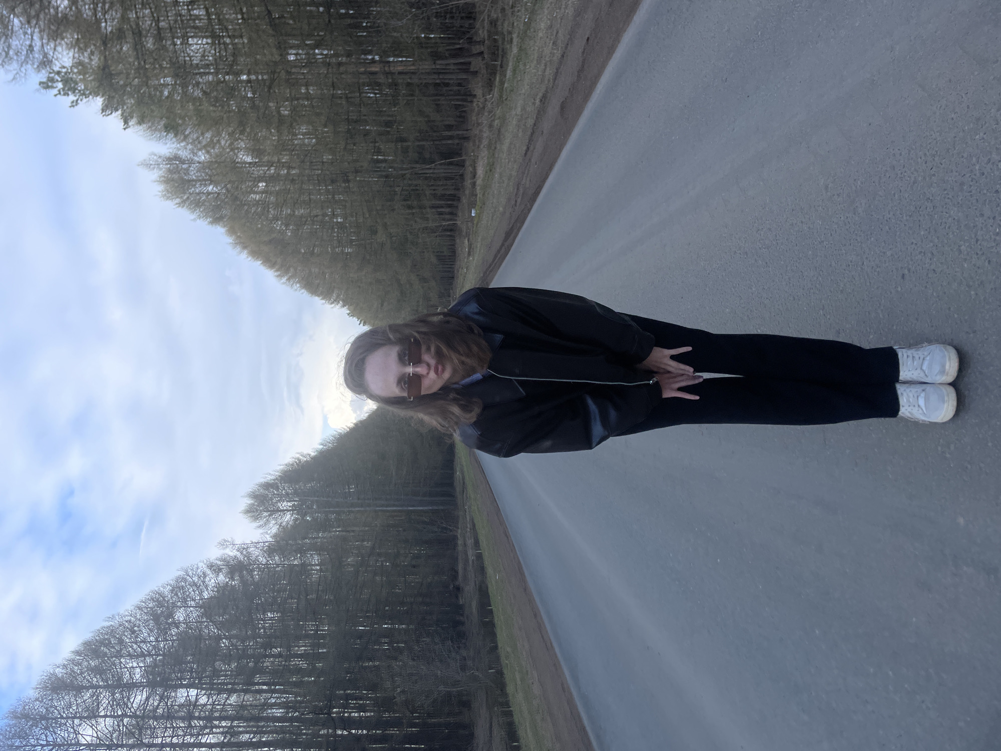 A person is positioned centrally on an asphalt road, looking directly at the camera. They wear dark sunglasses, a black faux leather jacket, dark trousers, and white sneakers. Their long, light brown hair frames their face, and their hands are clasped in front of them.
The road stretches into the distance, flanked on both sides by dense rows of tall, slender trees. Many of these trees appear bare or have minimal, light-colored foliage, suggesting a season of early spring or late autumn. The ground adjacent to the road is covered with dry grass and patches of dirt. The sky above is partly cloudy, displaying a mix of light blue and white clouds, indicating it is daytime. The setting is a rural or natural environment, consistent with a wooded area in Подшивалово, Russia. There is no visible text in the image.