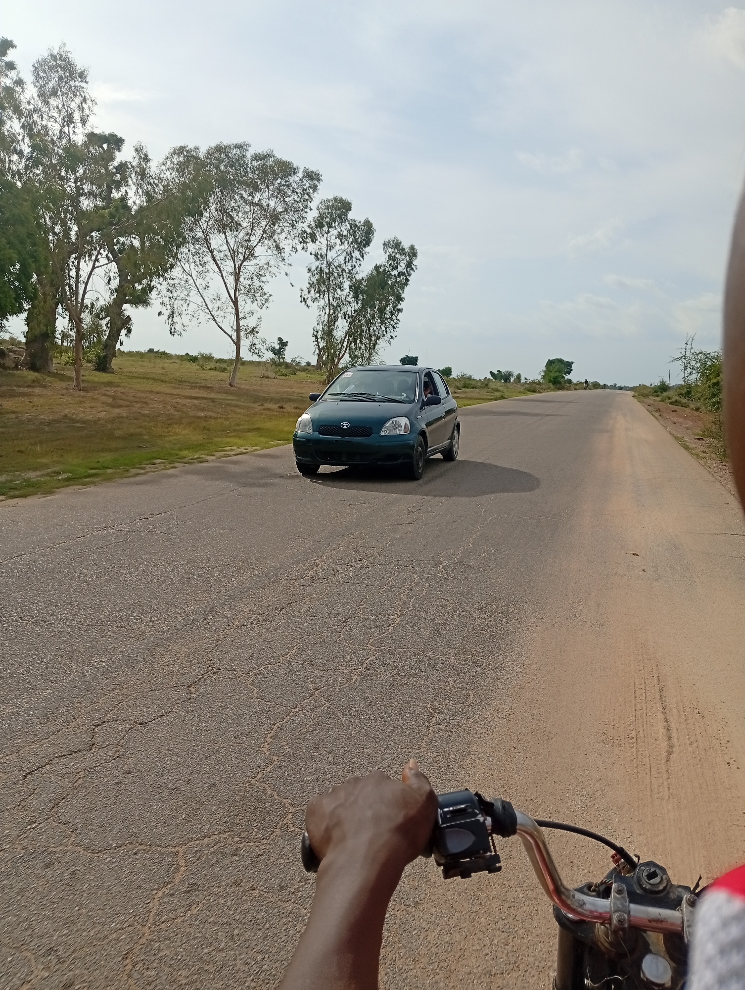 The image depicts a road in Shuwa, Nigeria, with a dark green Toyota Yaris driving on the asphalt road. The road is cracked and appears to be a two-lane highway. On the left side of the road, there are trees and dry grass. On the right side, there is a dirt shoulder and sparse vegetation. The sky is overcast with some clouds. In the foreground, the image is taken from the perspective of someone riding a motorcycle, with their hand on the handlebars. The rider's hand is visible, and they are wearing a gray sleeve on their arm. The motorcycle's handlebars, throttle, and brake lever are also visible. The scene appears to be a rural or semi-rural setting.