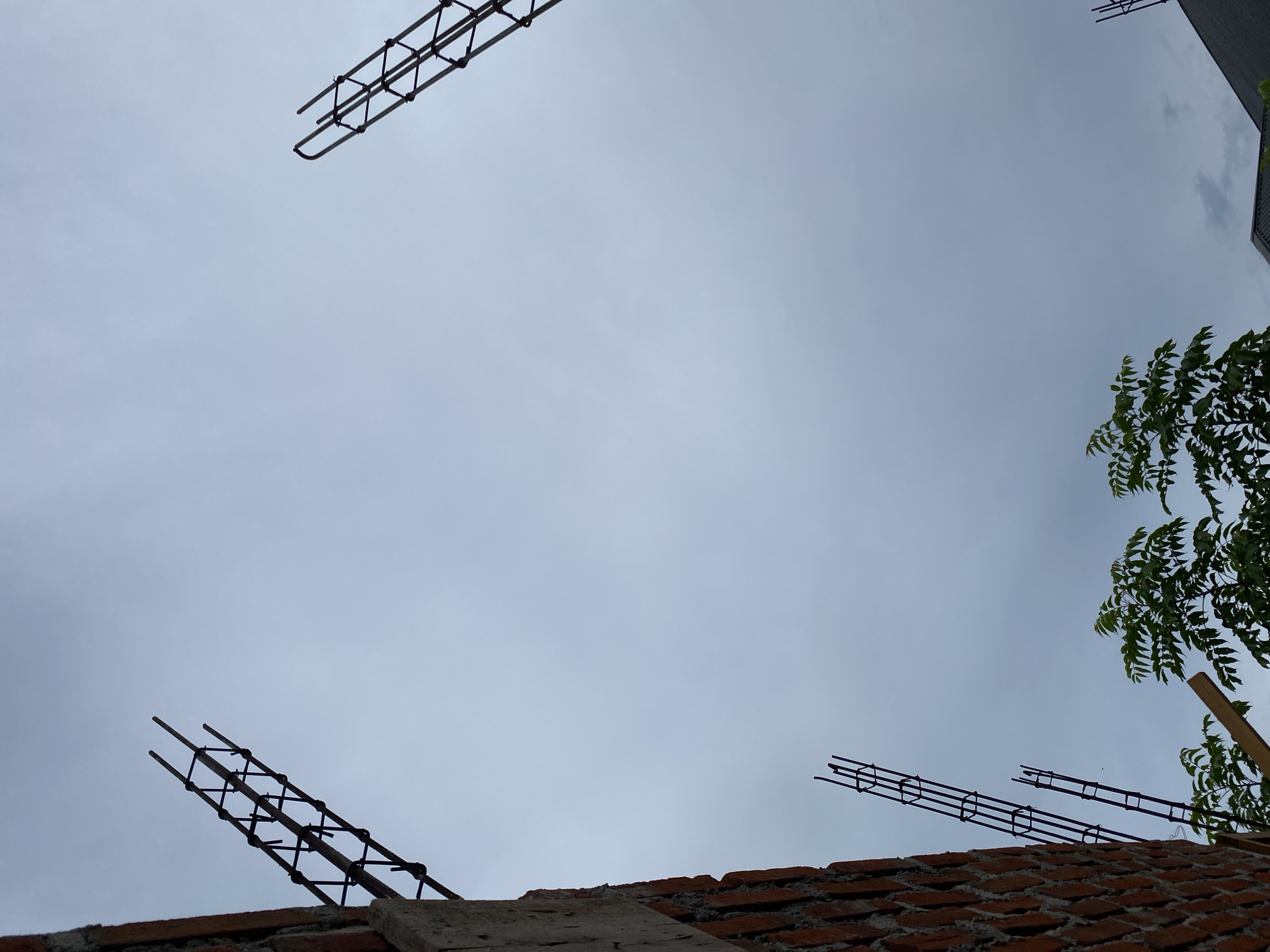 The image shows a construction site on an overcast day, with a cloudy sky. In the foreground on the left, a brick wall is partially visible. Extending upwards from the wall are reinforcing steel bars, forming a rebar cage, characteristic of a building under construction. Two more rebar cages are visible in the sky, one in the upper right and another further down on the left. In the bottom part of the image, the tops of green trees are visible, along with a wooden beam. The overall scene depicts an ongoing construction project in an outdoor setting, likely within the city of Banda Aceh, Indonesia, given the context provided. There are no people or animals present in the image.