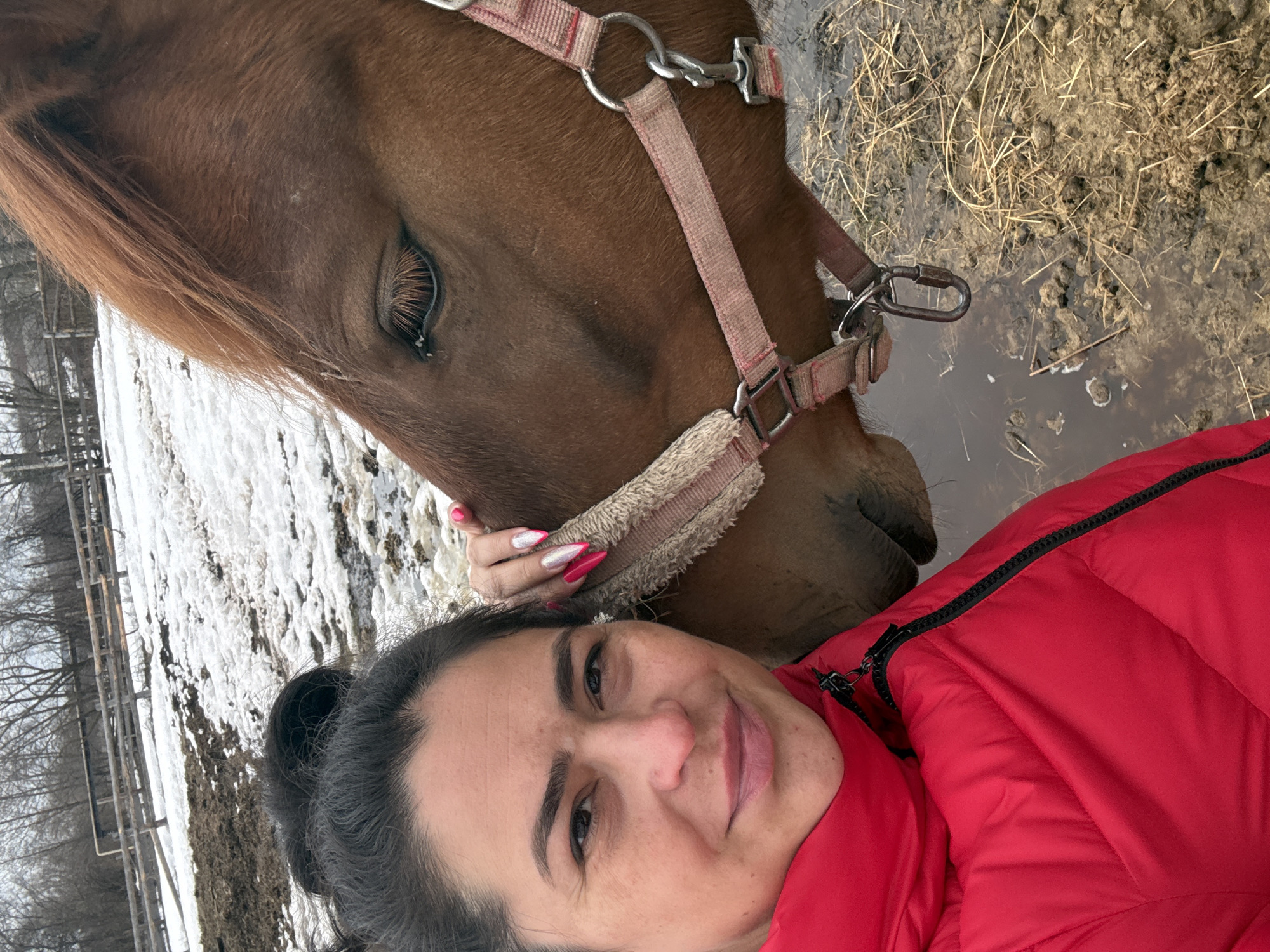 The image shows a selfie taken by a woman with dark hair pulled back, wearing a red puffer jacket. She is lying next to a chestnut horse with a pink and off-white halter. The background suggests a rural setting in the city of Кемуль, Russia, with patches of snow and muddy ground visible. The horse is partially submerged in a shallow puddle. The woman's manicured nails are visible as she touches the horse's muzzle. The setting appears to be an outdoor enclosure or pasture.