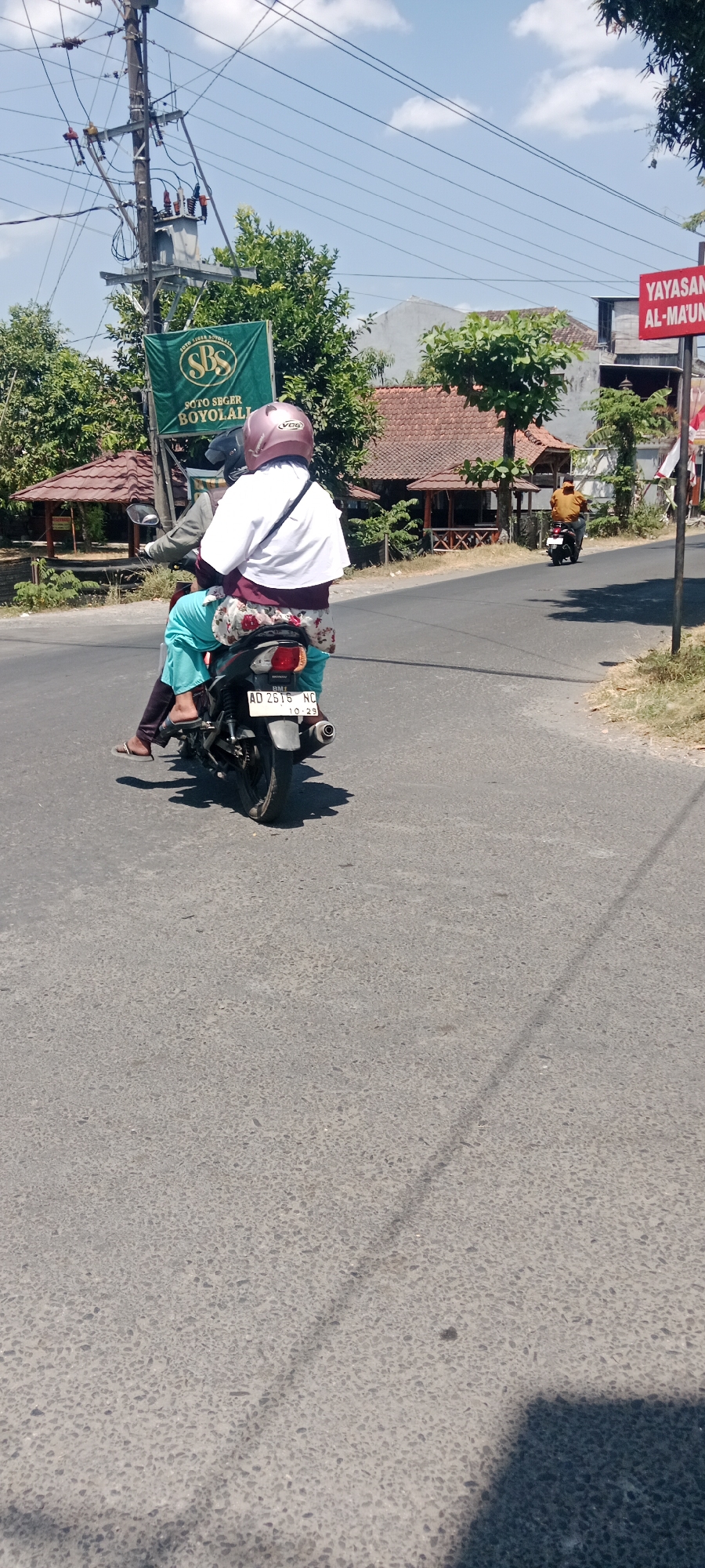 A full-shot, eye-level, outdoor, vertical medium-wide photograph captures two people riding a motorcycle on a paved road. The motorcycle is a dark color, possibly black, with a visible license plate reading "AD 2616 NC". The rider in the front is wearing a light-colored jacket over a dark shirt and a maroon helmet with a visor. The passenger behind is wearing a white outer garment over a dark top and patterned leggings, along with turquoise pants. They are both facing away from the camera, looking forward. The environment appears to be a sunny day in Maguwoharjo, Indonesia, with a clear blue sky and scattered white clouds. To the left of the road, a utility pole with electrical equipment and a green banner with the text "SOTO SEGER BOYOLALI" is visible. Behind the banner, lush green trees and buildings with terracotta roofs are present. Further down the road to the right, another motorcycle with a rider in an orange shirt is seen in the distance. A red sign with white text, possibly advertising "YAYASAN AL-MA'UN", is also visible on the right side of the frame. The road surface is asphalt with visible texture and some scattered debris. Shadows are cast on the road, indicating the presence of sunlight.