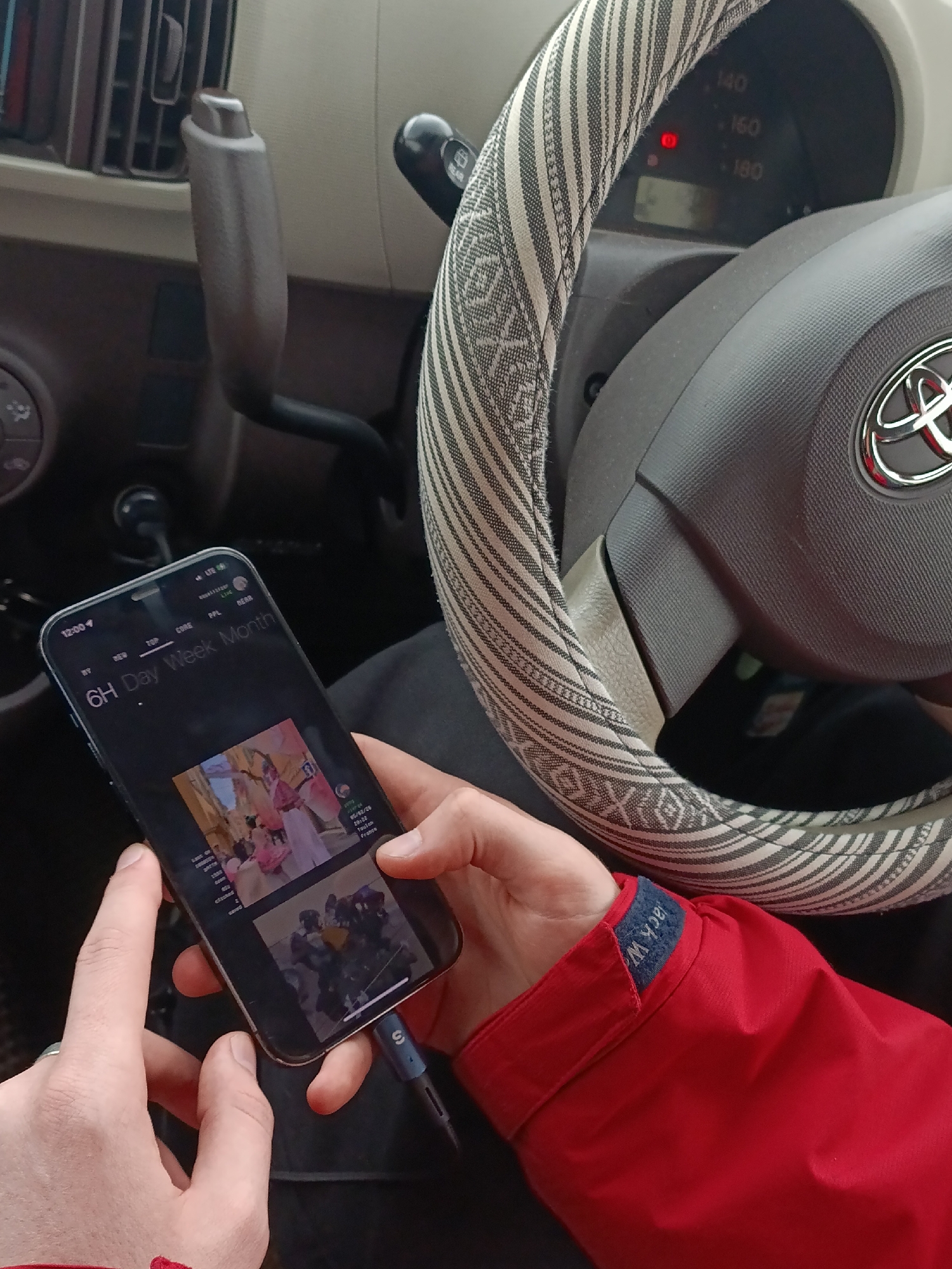 A person is in the driver's seat of a Toyota car. The phone screen displays a social media feed with a post. The phone is plugged into a charging cable. The car has a patterned steering wheel cover.
The OCR data suggests the location in the social media feed may be Toulon, France, not Krasnoyarsk, Russia.