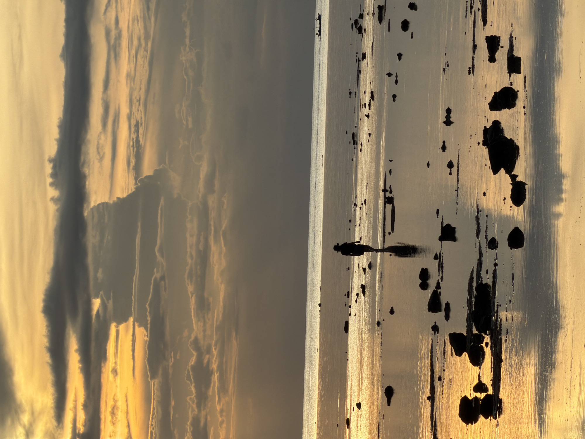 The image depicts a serene coastal scene at sunset in Baan Plai Laem, Thailand. The sky is a vibrant canvas of golden hues and dramatic grey clouds, with the setting sun casting a warm glow. In the foreground, a person wearing a hat walks along a wet, rocky shoreline, their silhouette stark against the reflective water. Scattered rocks dot the shallow water and the sand. In the distance, a small boat with two occupants can be seen on the calm sea. The overall atmosphere is one of tranquility and natural beauty.