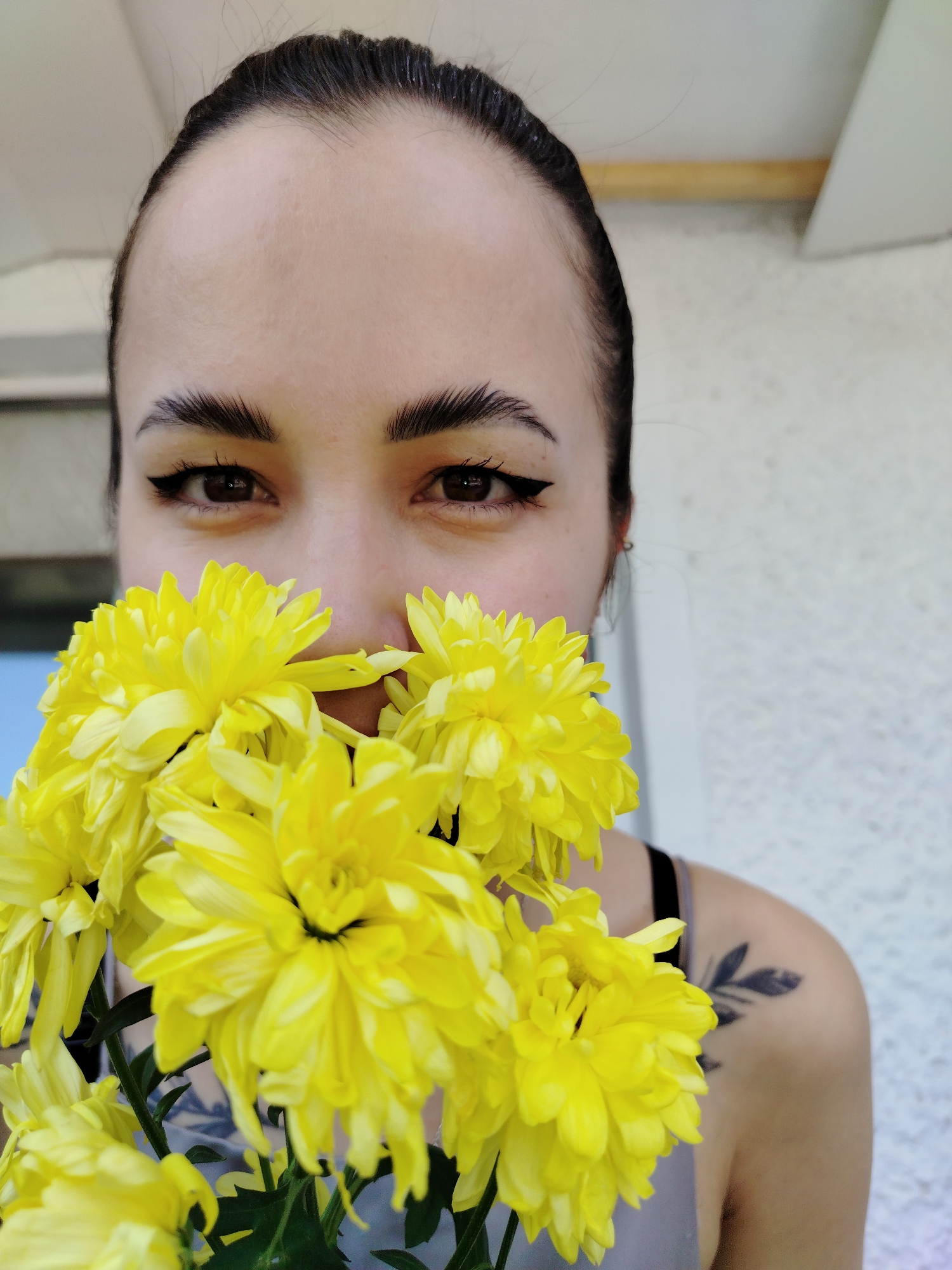 A woman is holding a bouquet of yellow chrysanthemums in front of her face, obscuring her nose and mouth. She is wearing black eyeliner and has her dark hair pulled back. A tattoo of a black leafy branch is visible on her right shoulder. The background appears to be an outdoor setting with a white wall or railing and glimpses of blue sky or building in the upper left. The location is Noviy, Russia.