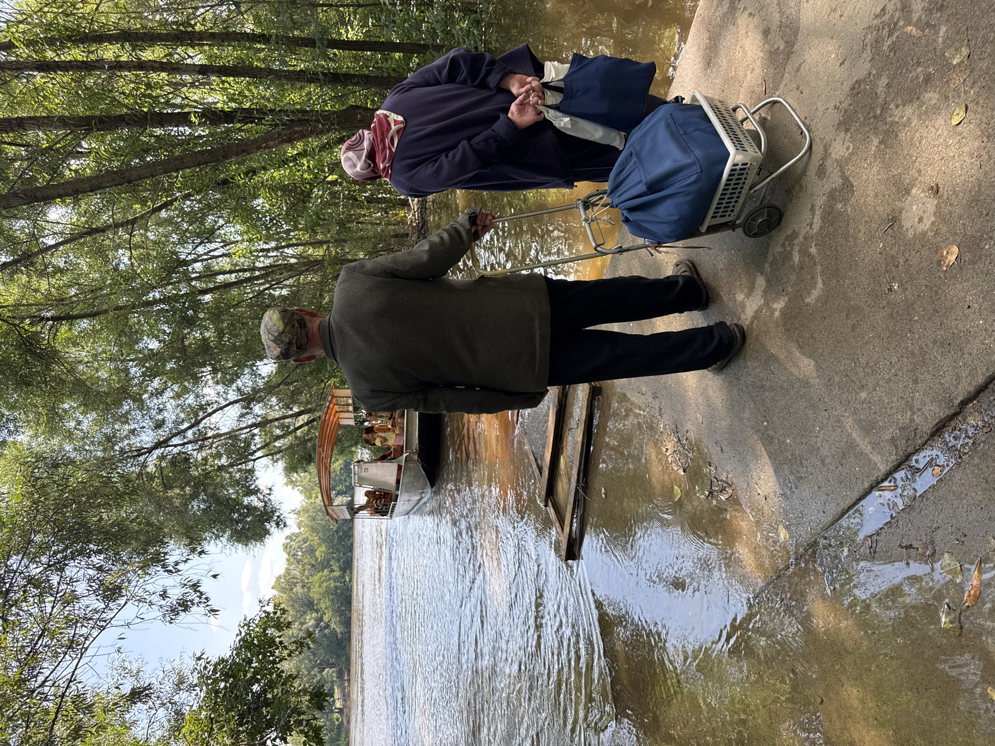 An elderly couple stands on a concrete ramp at the edge of a wide, muddy river, looking towards a small boat docked a short distance away. The man, wearing a camouflage cap and a dark jacket, holds a walking stick. The woman, in a headscarf and dark clothing, carries two shopping bags. A wheeled cart with a large blue bag sits between them. Trees line the opposite bank of the river, and the sky is clear and blue. The location is the Belaya River in Ufa, Russia, near a ferry crossing point.