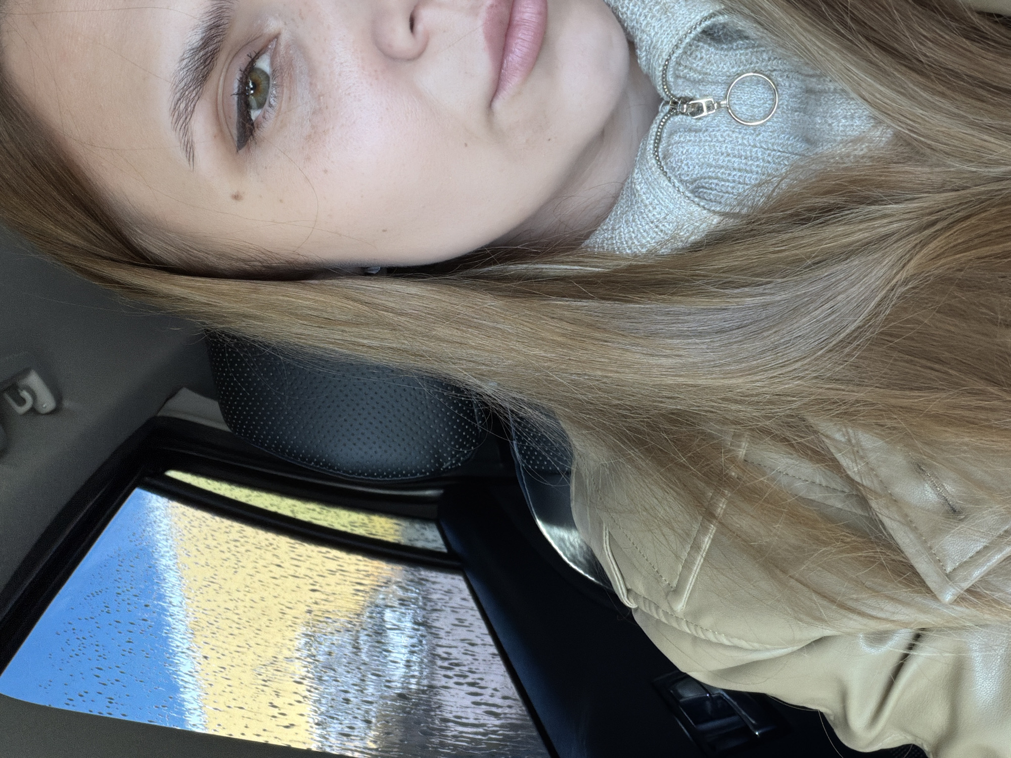 The image shows a selfie of a woman inside a car, likely taken during or after a rain shower, as evidenced by the water droplets on the window. She has fair skin, light brown hair, and green eyes. She is wearing a light-colored, possibly beige, jacket and a light gray knitted sweater with a zipper pull in the shape of a ring. The background visible through the car window is blurred, showing blue sky and reflective surfaces indicating a cityscape. While the city cannot be identified with certainty from the image, the request specifies Chaikovsky, Russia, as the location. Therefore, the image depicts a woman in a car in Chaikovsky, Russia.