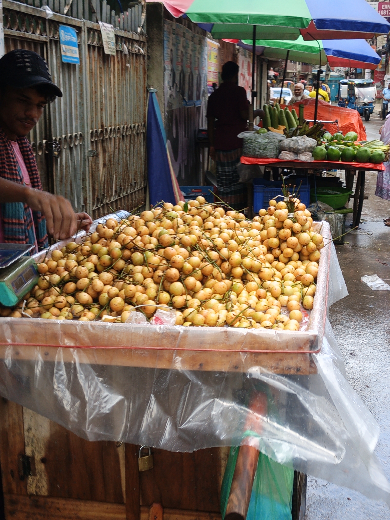 This is a close-up, eye-level, outdoor shot of a fruit stand in Dhaka, Bangladesh, under a slightly overcast sky. The foreground is dominated by a large wooden crate filled with a mound of small, yellow-orange fruits with stems. The crate is lined with clear plastic sheeting, and a red plastic tie is visible around the top edge. A metal scale with a teal-colored base is positioned to the left of the crate, with a person's hand reaching into the fruit. The person is wearing a pink shirt and a black baseball cap. Behind the fruit stand, a rustic metal fence with vertical bars and some peeling paint can be seen. A blue and white sign is affixed to the fence. To the right of the fruit stand, the ground is wet, suggesting recent rain, and other market stalls and people are partially visible in the background, with colorful umbrellas providing shade.