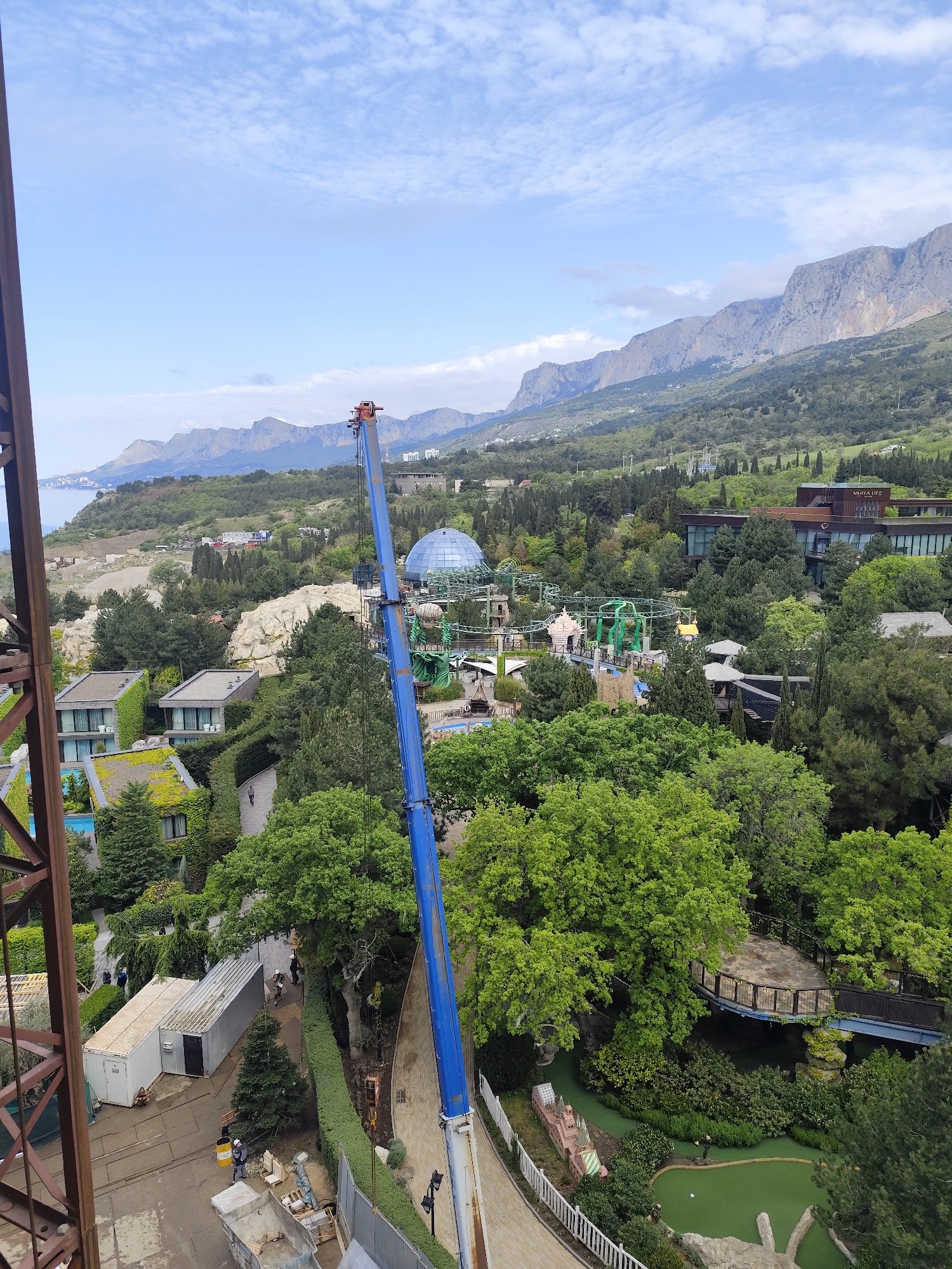 The image shows a view overlooking a landscape with a variety of elements. In the background, there is a mountain range stretching across the horizon under a sky with scattered clouds.
In the midground, there is a mix of vegetation, including various trees and green spaces. Distinct buildings are visible, including modern-style residences, a large blue dome structure, and a building with signage that reads "MRIYA LIFE." There is also a section that appears to be an amusement or theme park with winding roller coaster structures and pathways.
In the foreground, a construction crane with a blue boom occupies a significant portion of the frame. Below the crane, there are construction materials and a small building. A hedge-lined walkway is visible, and below that, there is a miniature golf course.