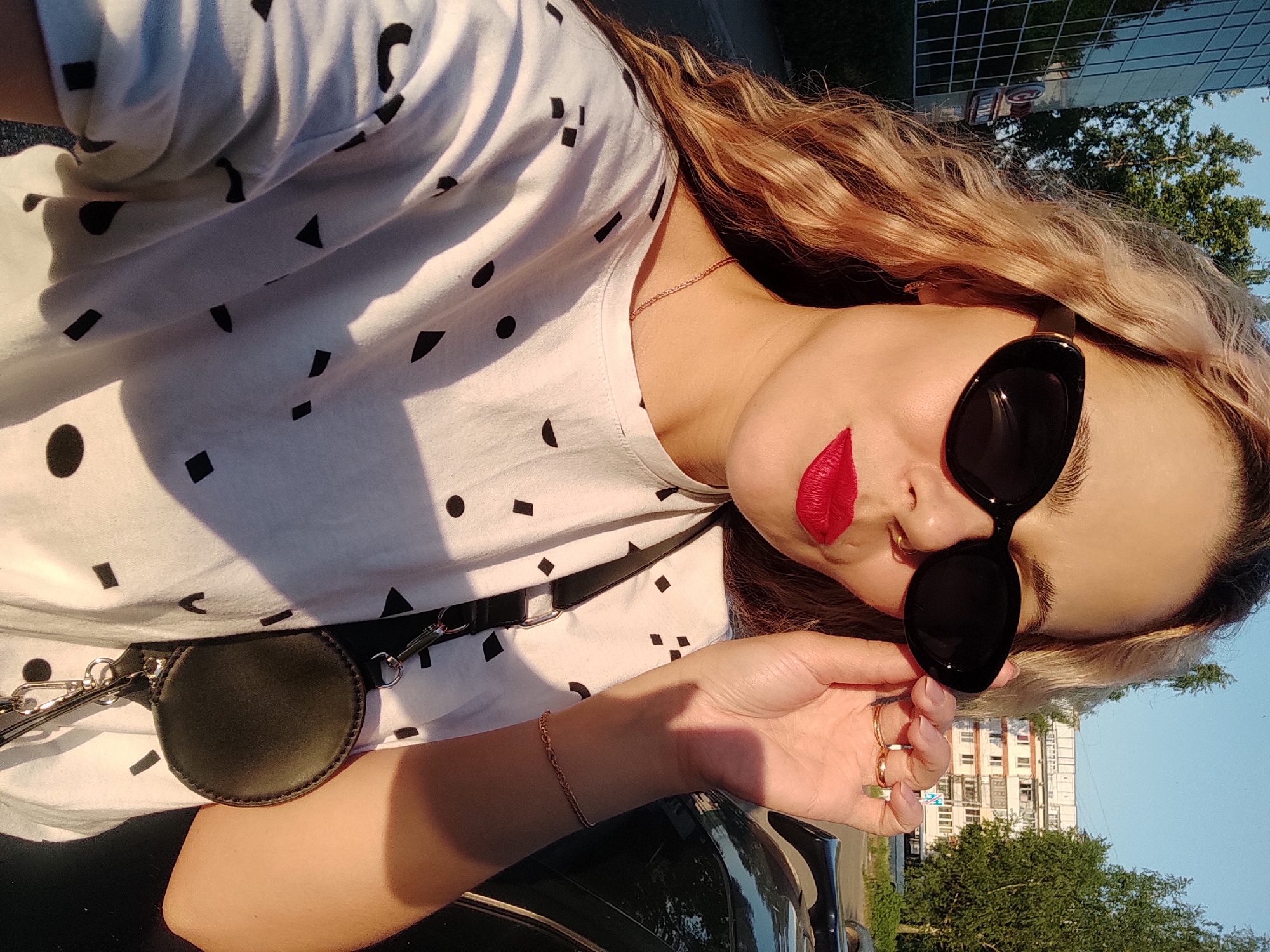 A woman poses, wearing sunglasses and red lipstick. She wears a white t-shirt with black shapes and a small, round bag on a strap. Her hair is long and wavy, and she wears gold jewelry. The background shows urban buildings and trees against a blue sky. The location is a city in an unknown location.