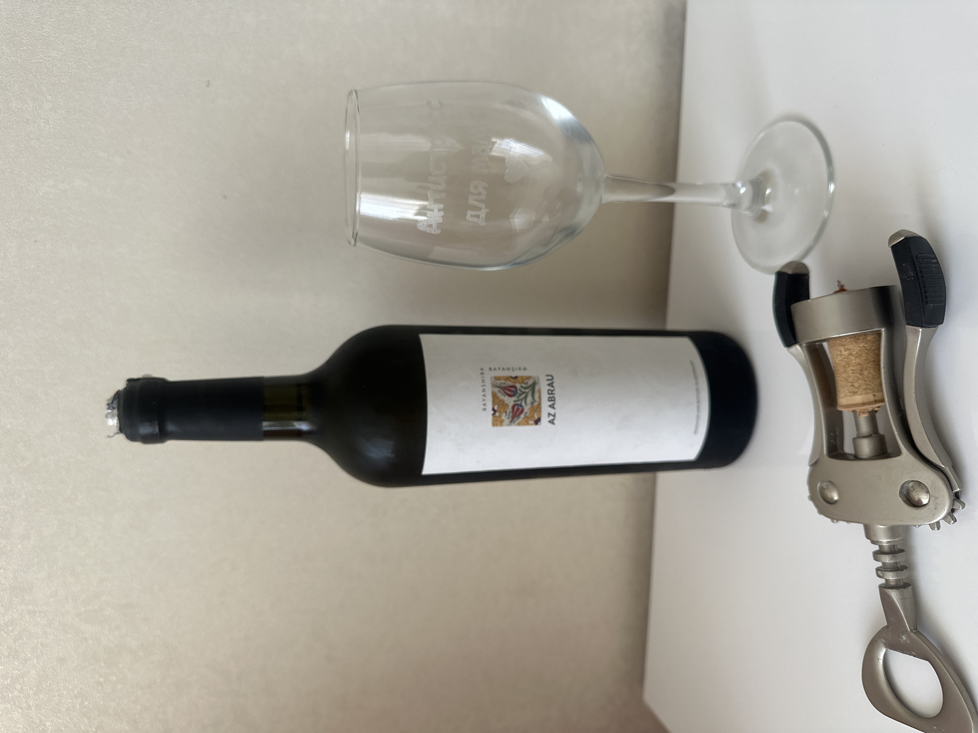 A still life image features a bottle of red wine, a wine glass, and a corkscrew placed on a white surface in front of a neutral-colored wall. The wine bottle, labeled "AZ ABRAU," is dark and has a black capsule around its neck, which appears slightly damaged at the top. The wine glass is clear and etched with text in a Cyrillic script and small heart designs. The corkscrew, made of metal, is positioned in the foreground with its cork screw partially inserted into a cork. The overall scene suggests an indoor setting, likely a home, in the city of Masazir, Azerbaijan.