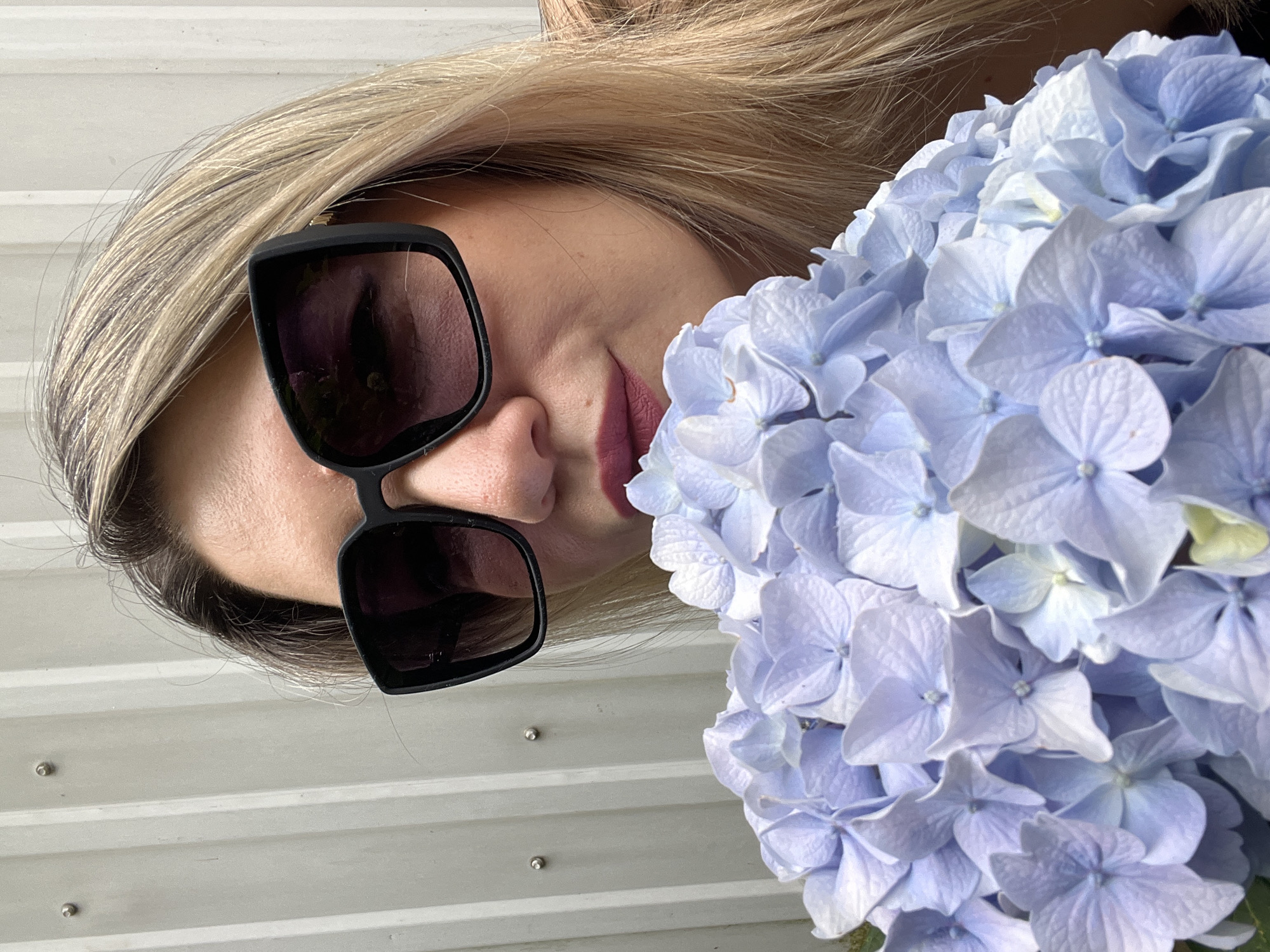 A woman with blonde hair is lying down, partially obscured by a bouquet of light blue hydrangeas. She is wearing black sunglasses that cover her eyes and her lips are painted with a matte pink lipstick. The background consists of corrugated white siding, suggesting an outdoor setting, possibly a balcony or patio. The lighting appears to be natural daylight, casting soft shadows. The overall mood is relaxed and serene, with the focus on the interplay between the woman's features and the vibrant flowers. No text is visible in the image.