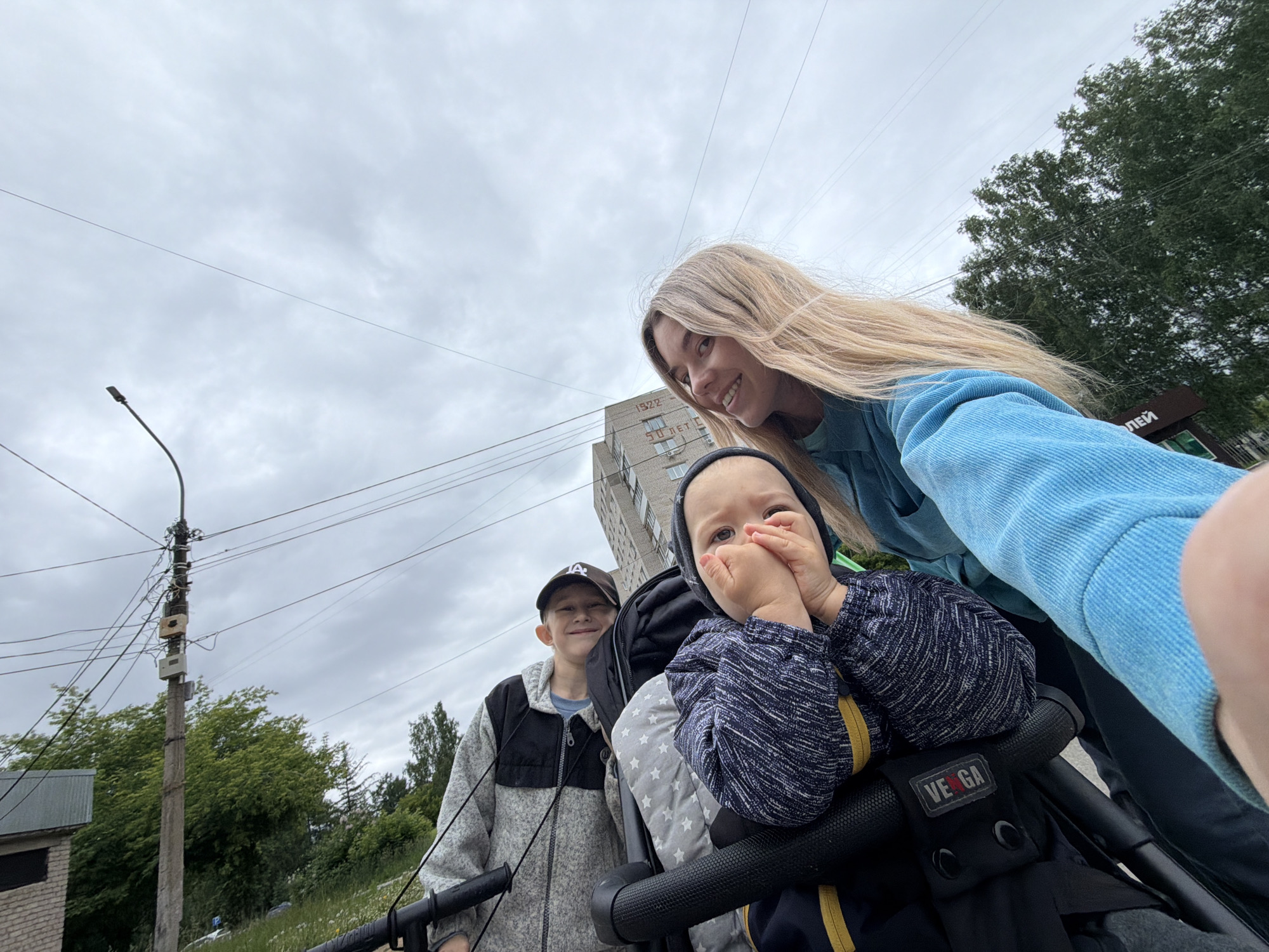 A low-angle, wide-view outdoor photograph taken in Chaikovsky, Russia, captures three individuals on an overcast day. In the foreground, a woman with long blonde hair, smiling, extends her left arm to hold the camera, partially obscuring the bottom right corner with her hand. She wears a blue corduroy jacket. To her left, slightly behind and lower, a baby or toddler with a hooded head is seated in a black stroller with a "VEGA" brand label and star-patterned grey padding. The child wears a dark blue top with light patterns and yellow stripes on the sleeves, with their hands covering their mouth. Behind the stroller and to the left, an older boy with short hair and a baseball cap (featuring a "LA" logo) stands, smiling at the camera. He is dressed in a grey and black zipped fleece jacket.
The background reveals a typical urban/suburban environment. To the left, a concrete utility pole heavily laden with numerous electrical wires and various grey and white electrical boxes stands prominently. Further left, a partial brick building is visible. In the middle-ground, a multi-story, light-colored brick apartment building features "1922" and "50 ЛЕТ" (50 Years) painted in red block letters on its facade. To the right of the building, a dense cluster of green deciduous trees is visible, with a sign partially reading "ЛЕЙ" (LEY) among them. The sky is largely overcast with diffuse grey clouds. Overhead, multiple thin wires crisscross the frame, emanating from utility poles.
