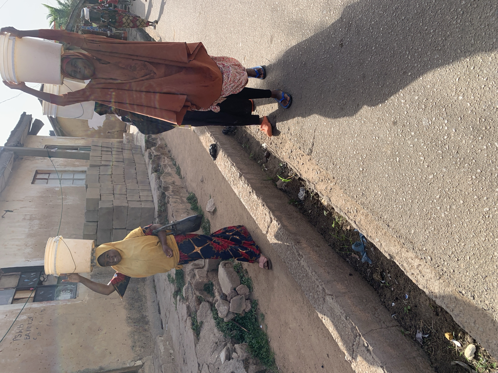 Two women carry water buckets on their heads in Jos, Nigeria. One woman wears a yellow head covering and floral skirt, while the other wears a brown head covering and floral top. They are walking near a street and buildings made of concrete blocks.