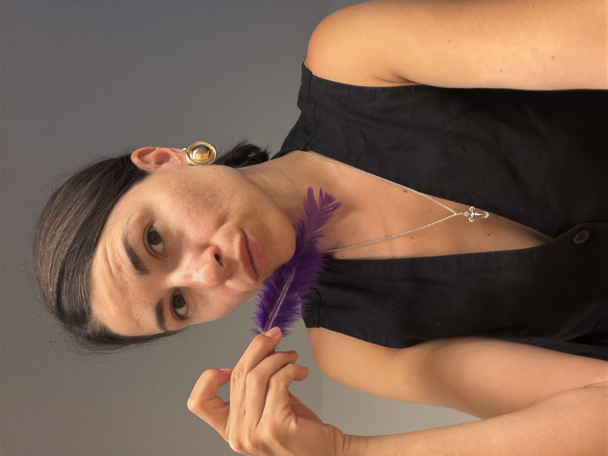 The image is a portrait of a person, likely a woman, with dark hair pulled back, looking directly at the viewer with a neutral to slightly curious expression. Her head is tilted to her left, and the image is oriented as if she is lying down or the camera was angled, with her head towards the left side of the frame. She has fair to medium-toned skin with some visible freckles across her face and body.
She is wearing a plain black sleeveless top or dress. On her left ear (viewer's right), she has a prominent, shiny gold-colored circular earring. A thin silver chain necklace with a small, intricate silver cross pendant rests against her chest.
With her right hand, she delicately holds a vibrant, fluffy purple feather, positioning it near her collarbone and neck. Her fingers are visible, gently gripping the base of the feather.
The background is a solid, unadorned gray wall or surface, providing a simple and clean backdrop. The lighting is soft and even, without harsh shadows, suggesting an indoor setting or a controlled studio environment. There is no visible text in the image. This portrait was taken in Budva, Montenegro.