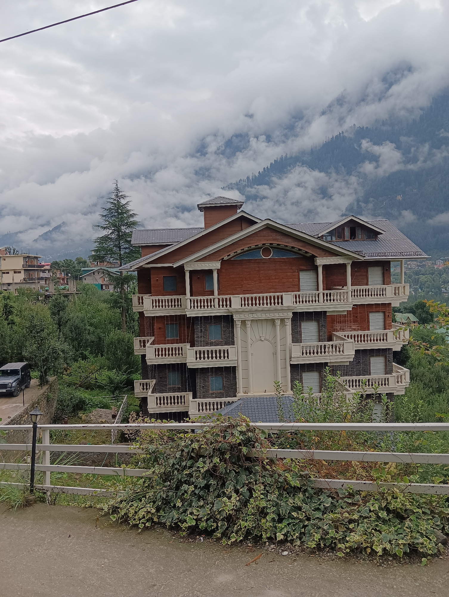 In Chhyal, India, a multi-story wooden building with decorative white balconies stands prominently against a backdrop of cloudy mountains. The building features a dark, textured roof and several windows with white frames. A dark-colored SUV is parked on a dirt road to the left of the building, surrounded by lush green trees. A white metal fence runs horizontally across the foreground, partially obscured by dense green foliage. The sky above is overcast with thick clouds, obscuring the mountain peaks.