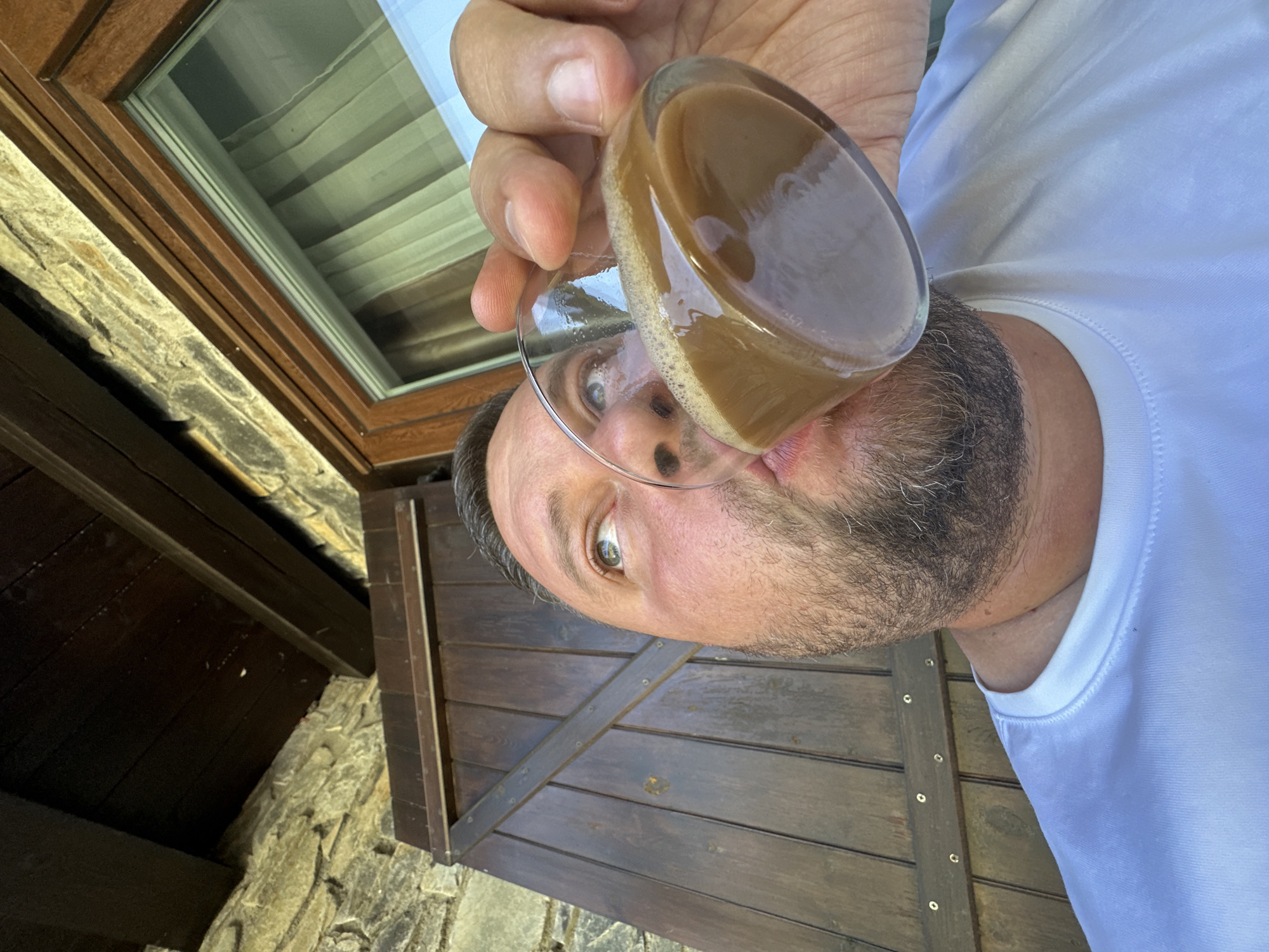 A dynamically angled portrait captures a man with a beard and short dark hair, wearing a white t-shirt. His head is tilted sharply to the side, making it appear as if he's looking up or the camera is positioned overhead. He holds a clear glass filled with a light brown, frothy beverage, likely coffee or a milky drink, which is tilted towards his mouth, suggesting he is about to drink or has just taken a sip. His eyes are wide open, fixed directly at the camera, conveying a surprised or playful expression.
The background on the upper left features a wooden-framed window with reflections of what seem to be horizontal blinds or curtains. Adjacent to the window, a textured stone wall is visible, suggesting traditional masonry. The lower right background shows a rustic wooden slatted structure, possibly a shutter or a door, complete with visible metal fasteners. Below this structure, stone pavers or rough ground can be discerned. The overall lighting is bright and natural, indicating daytime. The architectural elements like the stone work and wooden structures, along with the outdoor setting, are consistent with the provided location context of Encamp, Andorra. There is no visible text in the image.