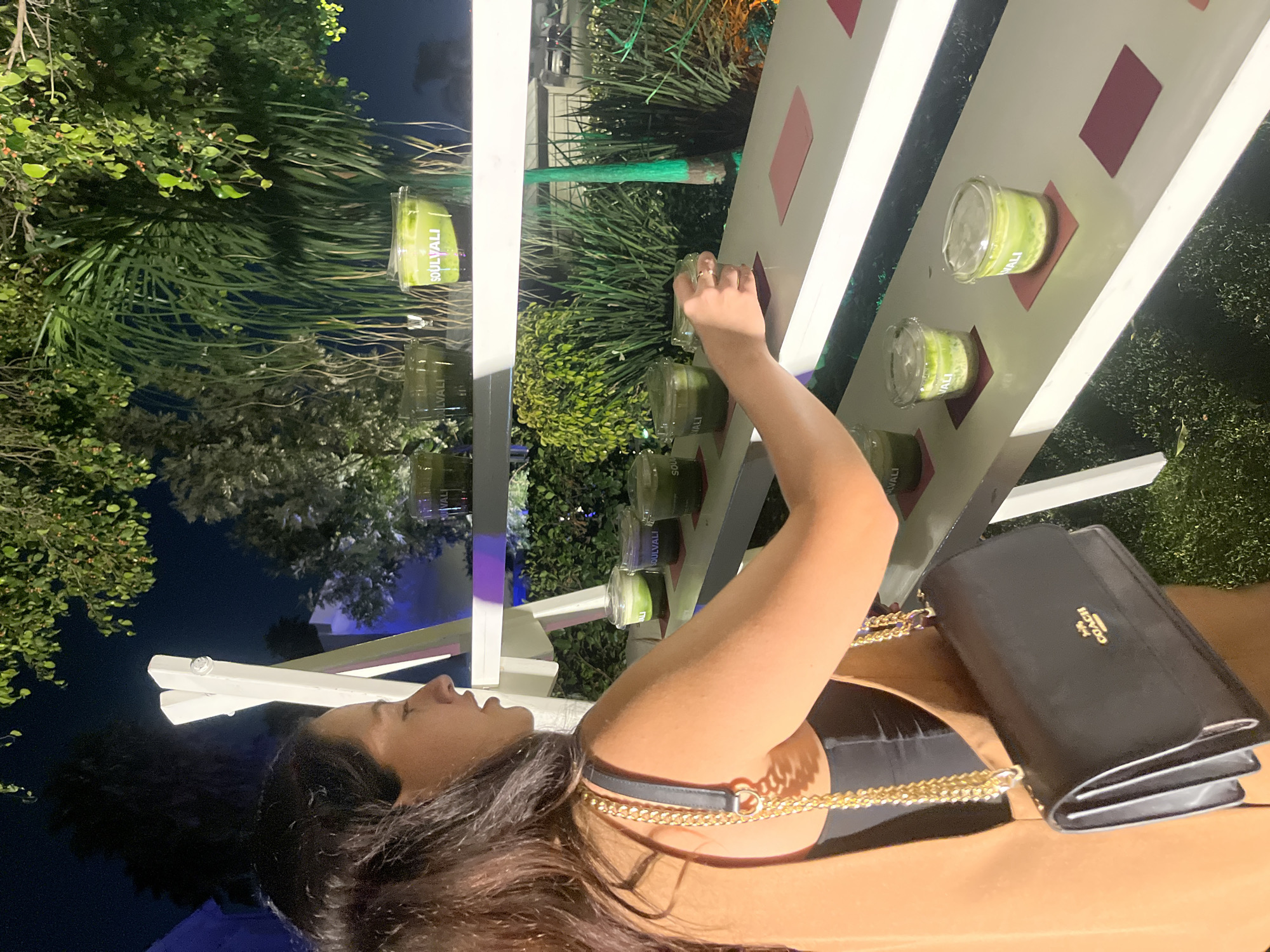 The image shows a woman standing in San Bernardino Tlaxcalancingo, Mexico, beside a white structure displaying small containers labeled "SOULYALI." The containers appear to hold a green substance. The woman has dark hair and is wearing a black top and a tan jacket or dress. A black Coach purse hangs on her shoulder, suspended by a gold chain strap. Vegetation is visible in the background.