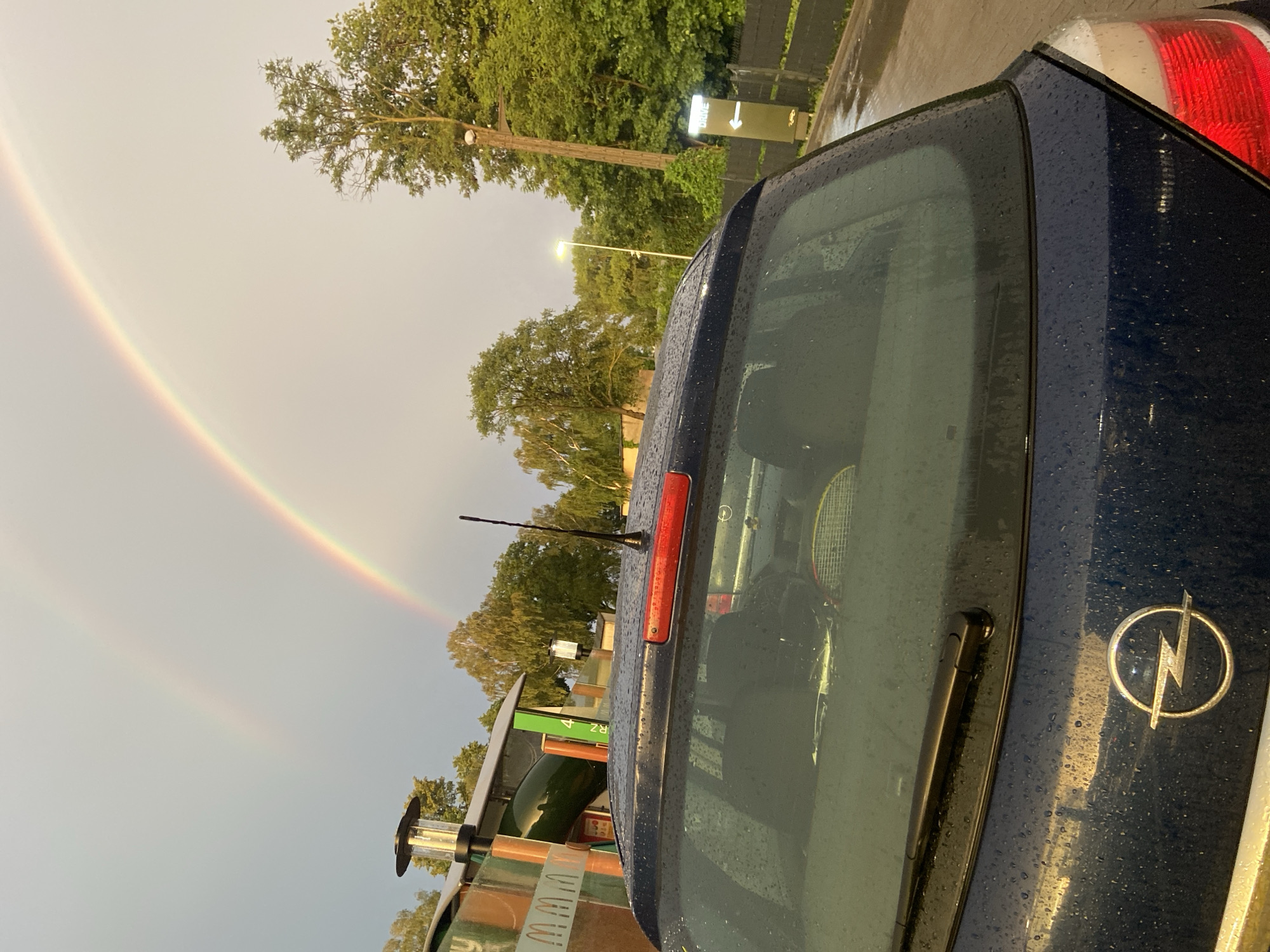 The image shows the back of a dark blue car parked outdoors. Raindrops are visible on the car's surface. Through the rear window, a tennis racket can be seen inside the car. An Opel logo is visible on the trunk of the car. To the left of the car, a rainbow arcs across a bright, overcast sky. Trees and some structures are visible in the background, including a sign with the word "DRIVE" and an arrow pointing to the right. The scene appears to be a drive-through or service area. The overall lighting suggests it is daytime, possibly late afternoon or early morning given the soft light and overcast conditions. The presence of rain and a rainbow indicates recent or ongoing precipitation. The location context is Bolesławiec, Poland.