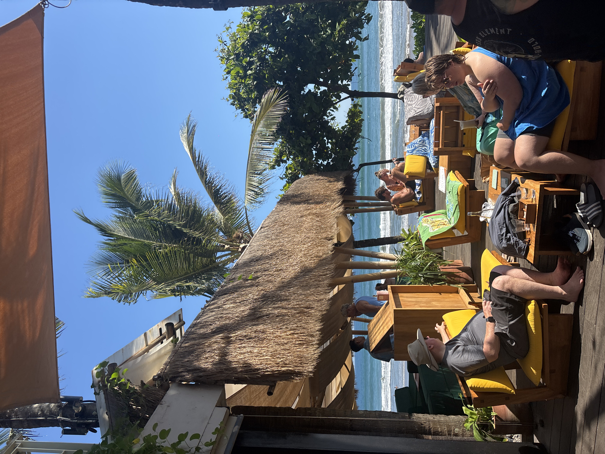 This image depicts a beachfront dining or lounge area in North Kuta, Indonesia. The scene is outdoors, with a thatched roof structure providing shade. Several people are seated on bright yellow lounge chairs, some facing the ocean, others interacting with each other. In the foreground, a man wearing a grey t-shirt and shorts, and a wide-brimmed hat, is seated on a yellow chair, looking to his right. To his right, a younger person in a blue garment is also seated on a yellow chair, looking to their left. Further back, other patrons are visible enjoying drinks and conversation. Palm trees and lush green foliage are present, with the ocean and waves visible in the background under a clear blue sky. The flooring appears to be made of wood.