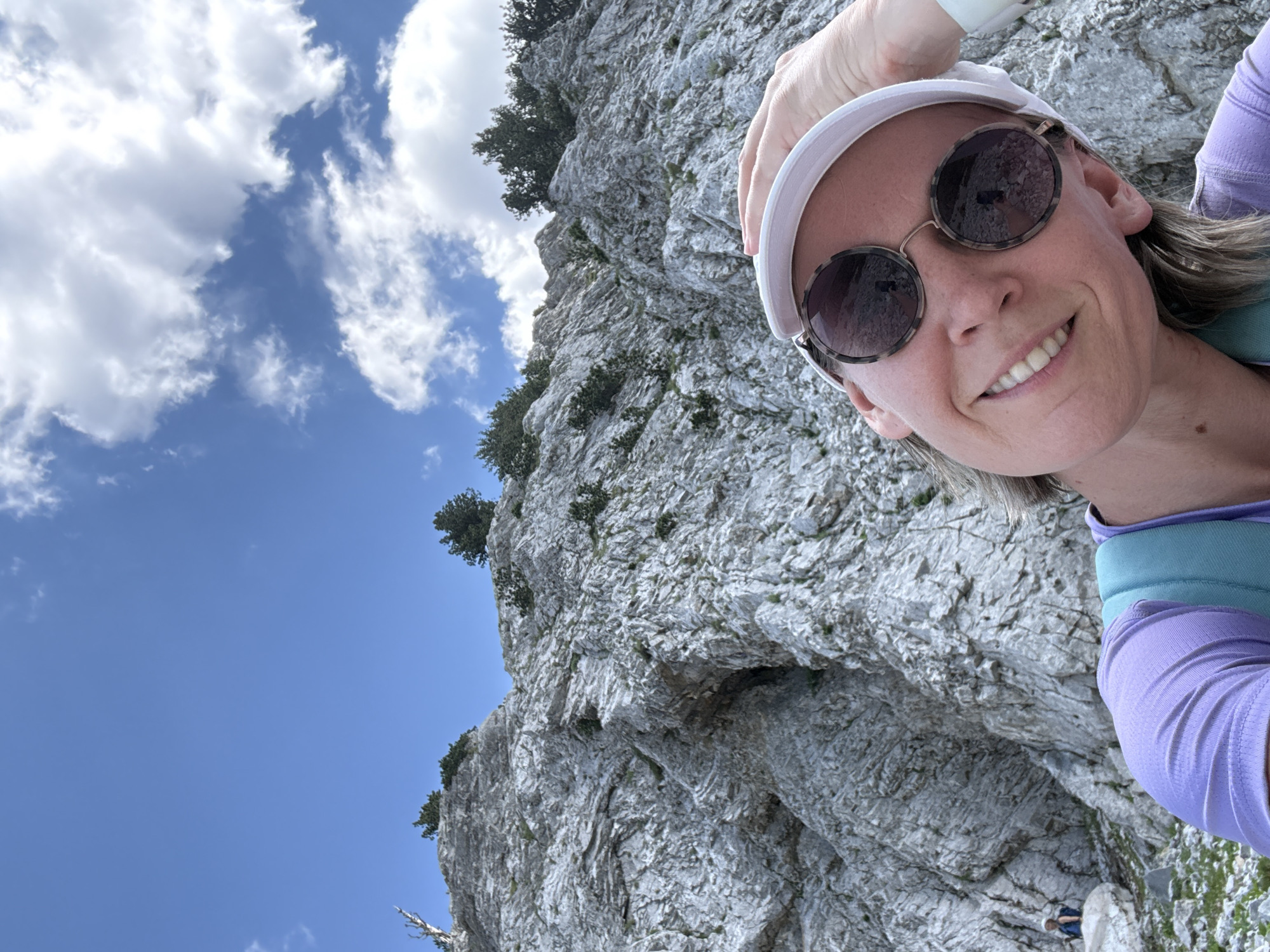 A woman, wearing a white cap, sunglasses, and a purple shirt with a turquoise backpack, takes a selfie outdoors. She is smiling and her right hand is on her head, pushing her cap back. In the background, a rocky mountain face with sparse green vegetation rises towards a bright blue sky dotted with white clouds. A person in a blue shirt and white hat is visible sitting on a rock in the distance on the left side of the image. The scene appears to be in a mountainous region, possibly Katun Vulića, Montenegro, given the landscape.